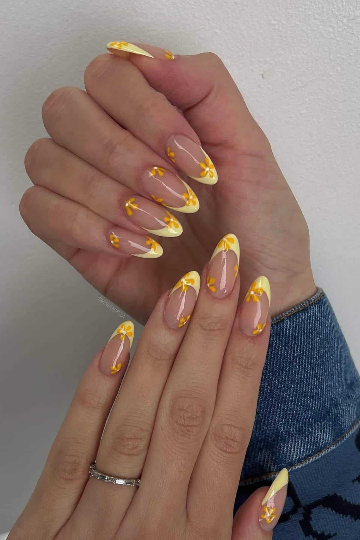 Hands with long almond-shaped nails featuring yellow French tips and small yellow flower designs. One hand rests on top of the other against a light background.