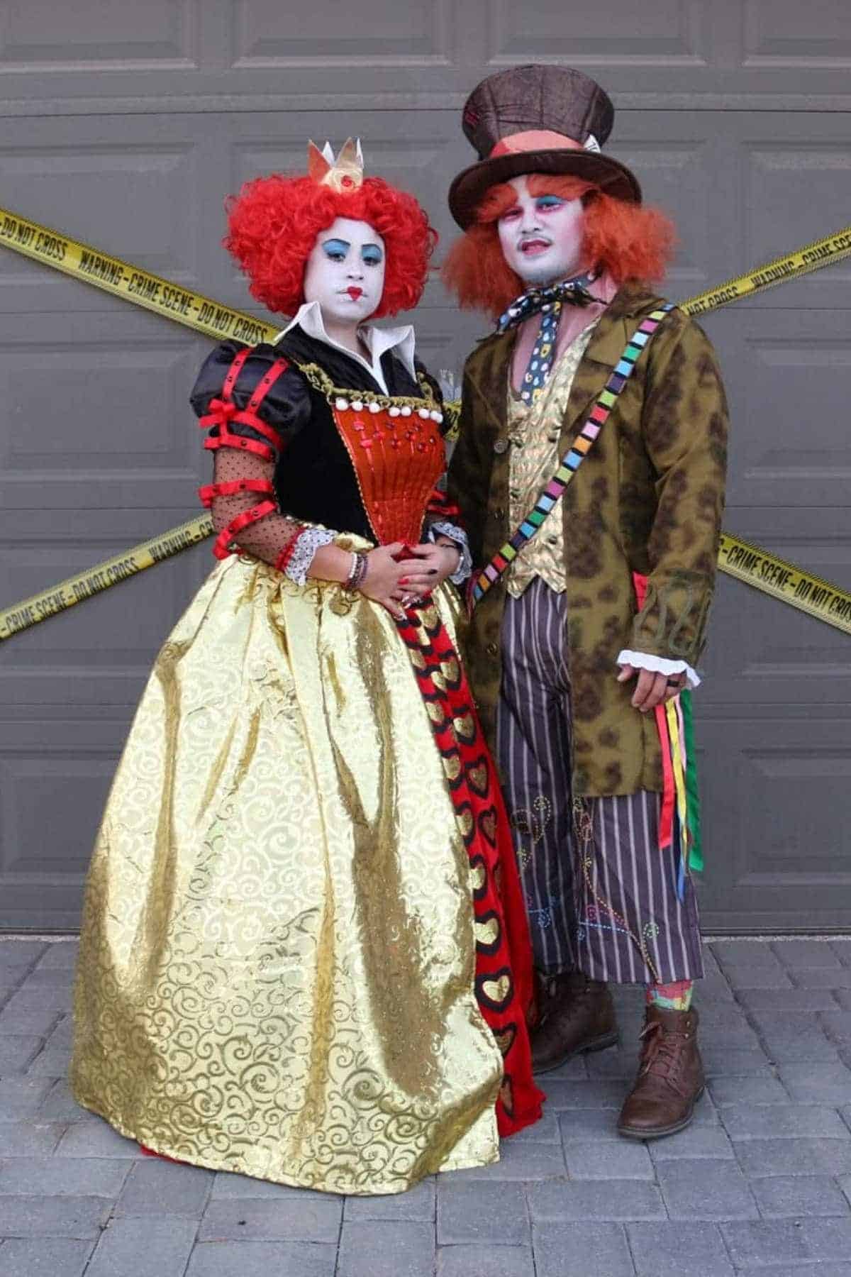 Two people dressed as the Queen of Hearts and the Mad Hatter from Alice in Wonderland stand in front of a garage door with "Do Not Cross" tape in the background.