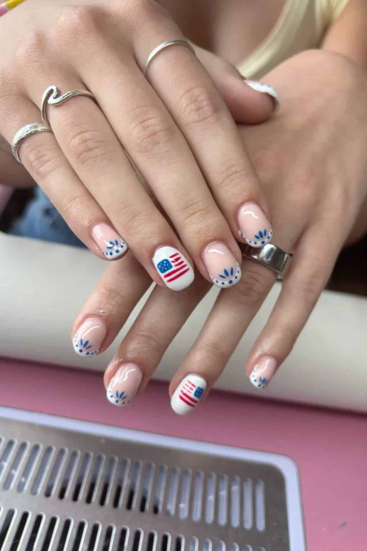 Hands with manicured nails featuring blue and white flower designs and American flag accents on clear polish. Person wears silver rings and a cream top.