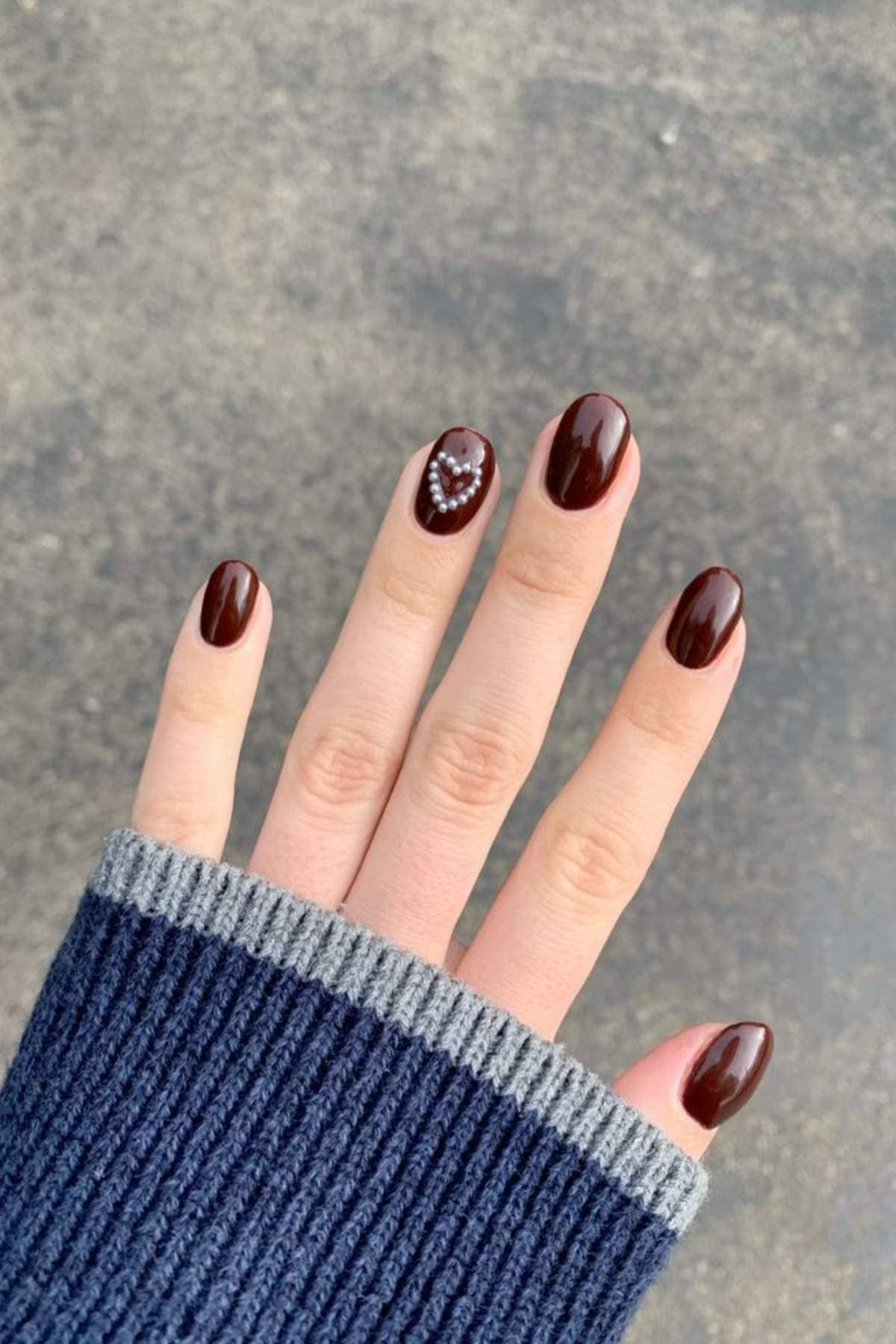 A hand with dark brown oval-shaped nails, one featuring a small heart design made of rhinestones, is shown against a gray background. The person is wearing a navy and gray sweater.