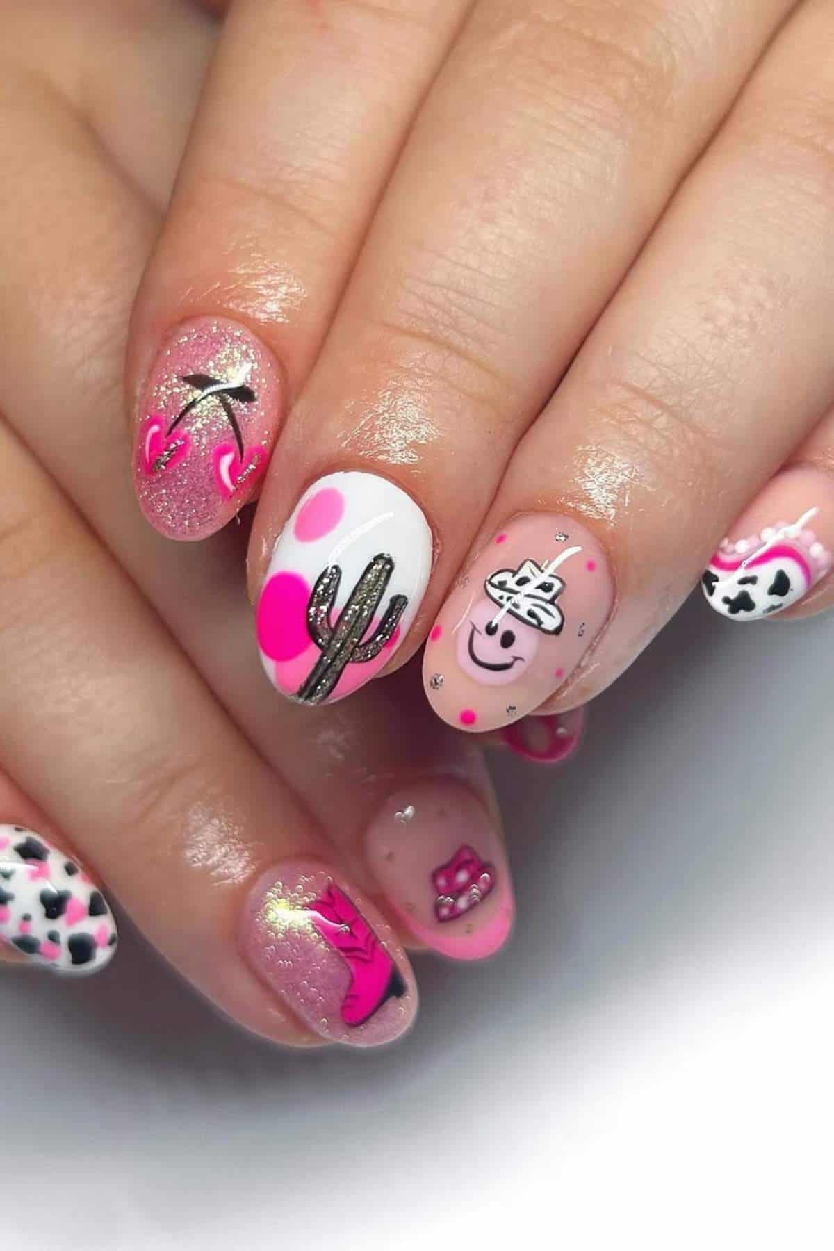 A hand with manicured nails featuring pink, white, and black designs, including a cactus, smiley face, hearts, crowns, and cow print patterns.