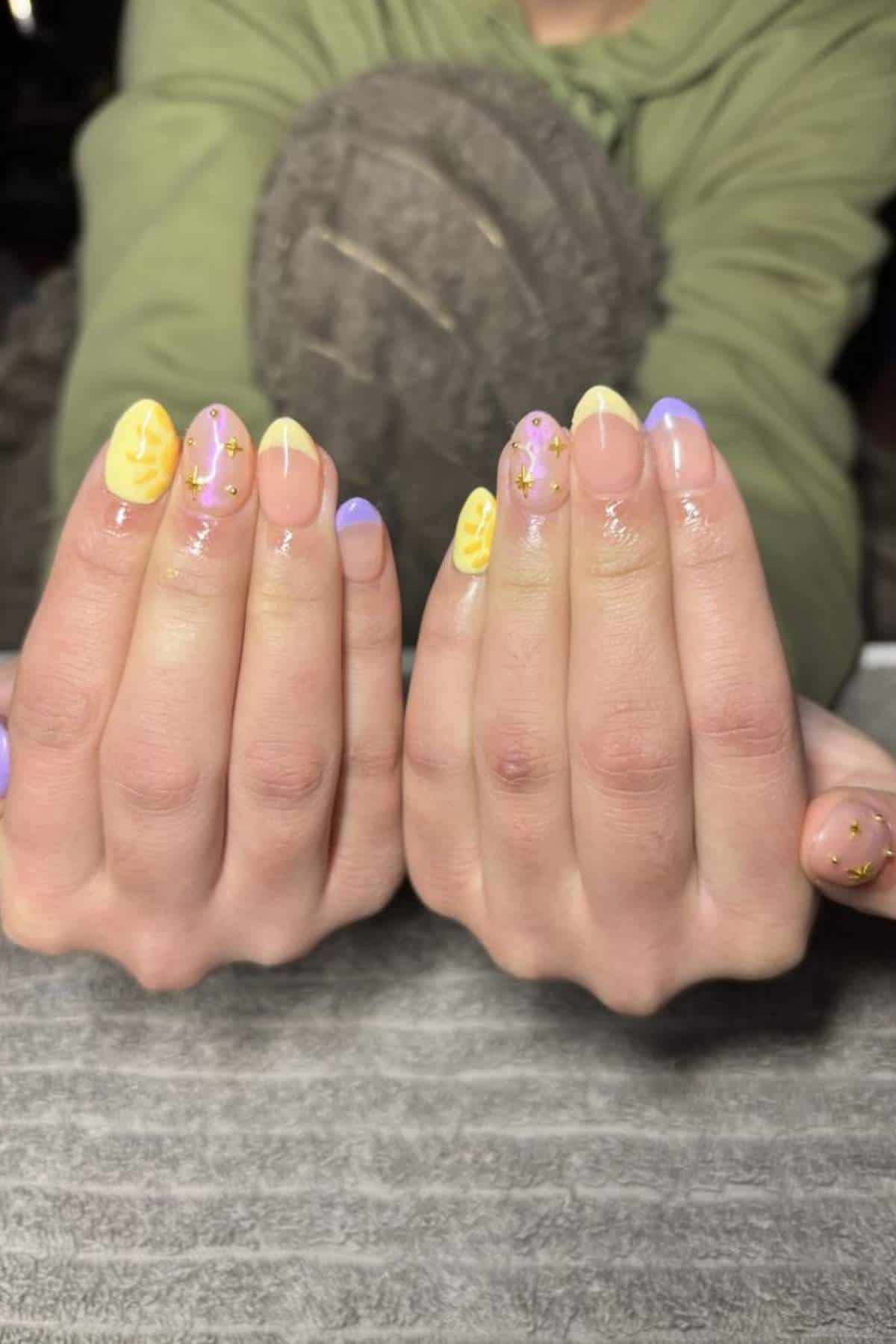 Close-up of hands with pastel yellow and purple manicure, featuring leaf designs and small gold accents on each nail.