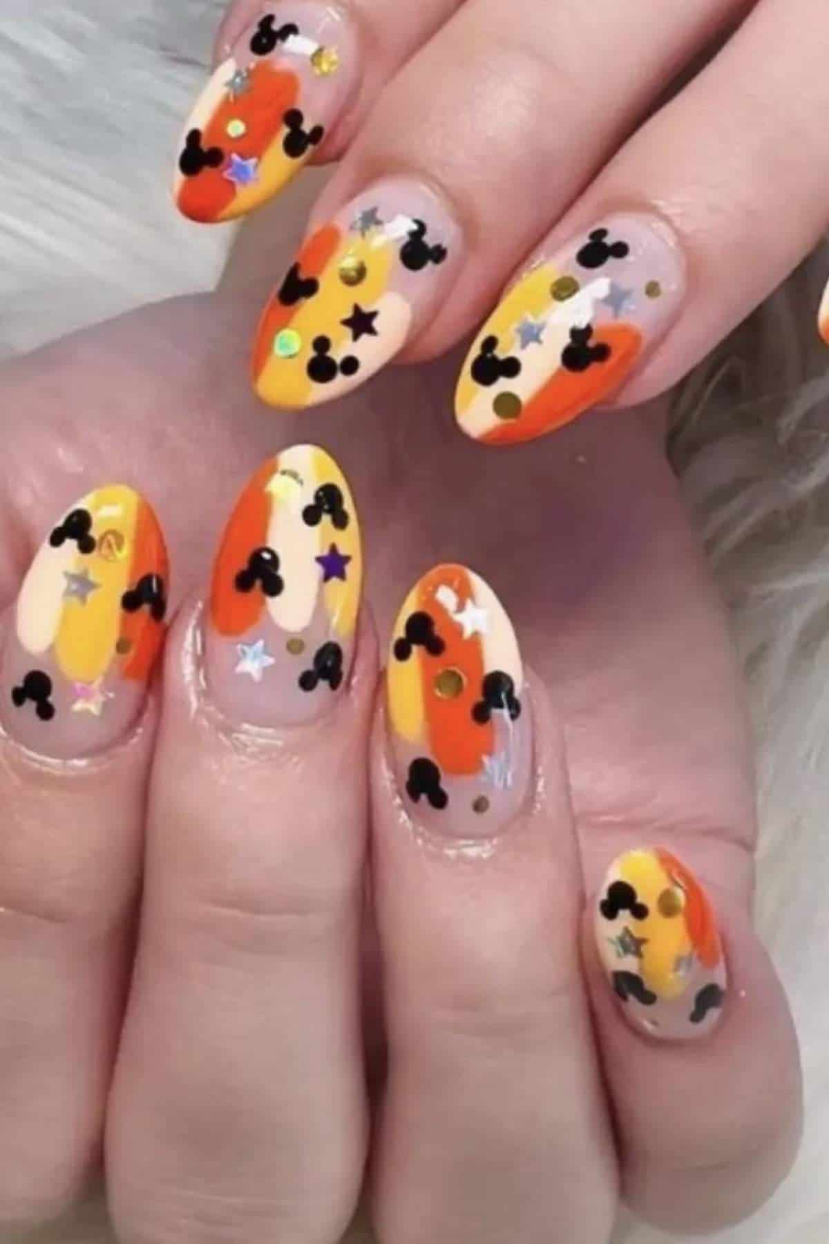 Close-up of hands with almond-shaped nails painted in yellow, orange, and nude tones, decorated with black Mickey Mouse shapes, stars, dots, and sequins.