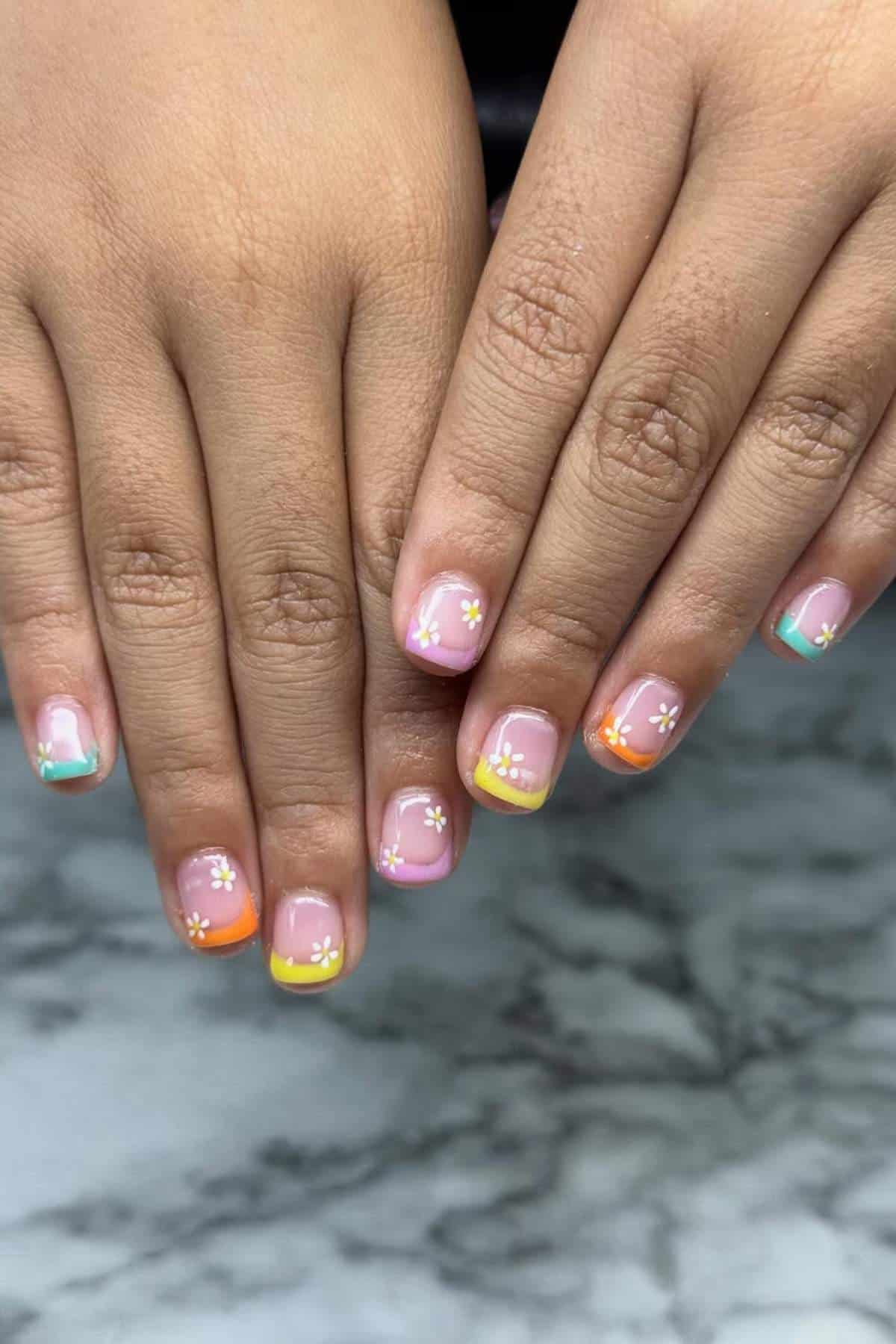 Two hands with short nails featuring multicolored French tips and small white flower designs, displayed on a marble surface.