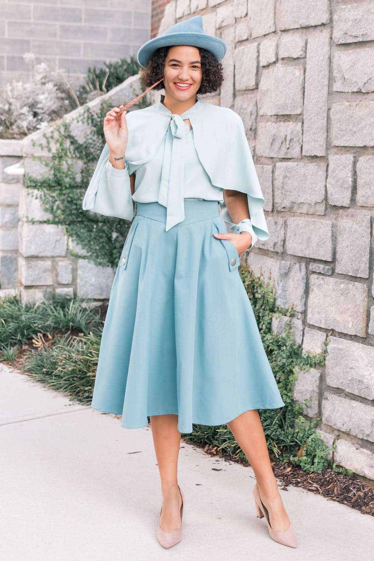 A woman stands outdoors in front of a stone wall, wearing a light blue cape, matching skirt, blouse with a bow, wide-brim hat, and nude heels.