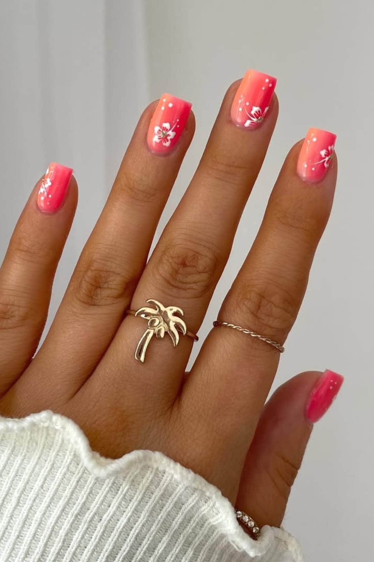 A hand with ombre pink and orange square-shaped nails, decorated with small white floral designs, wearing a palm tree ring and a thin band.