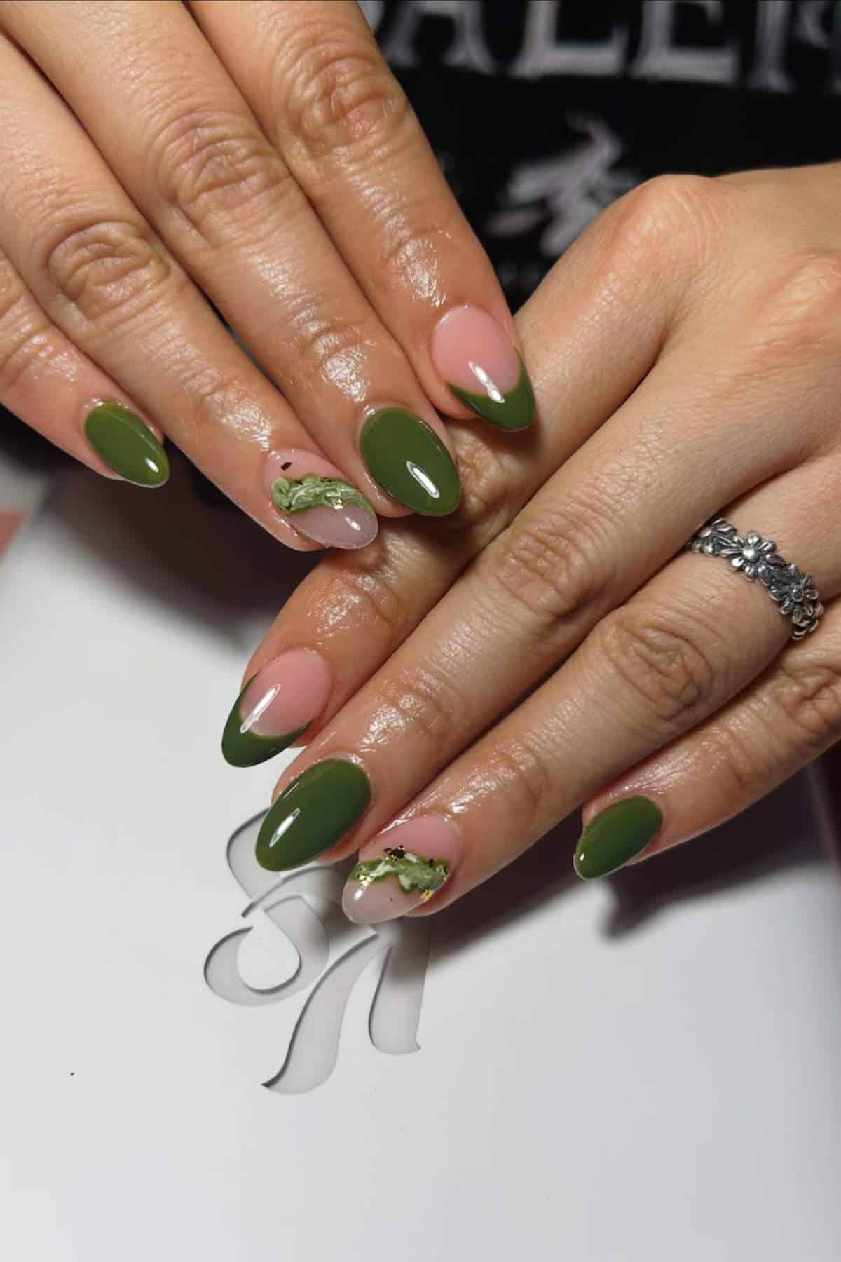 Hands with almond-shaped nails featuring green polish, pink base with green French tips, and gold foil accents on a few nails. A silver floral ring is worn on one finger.