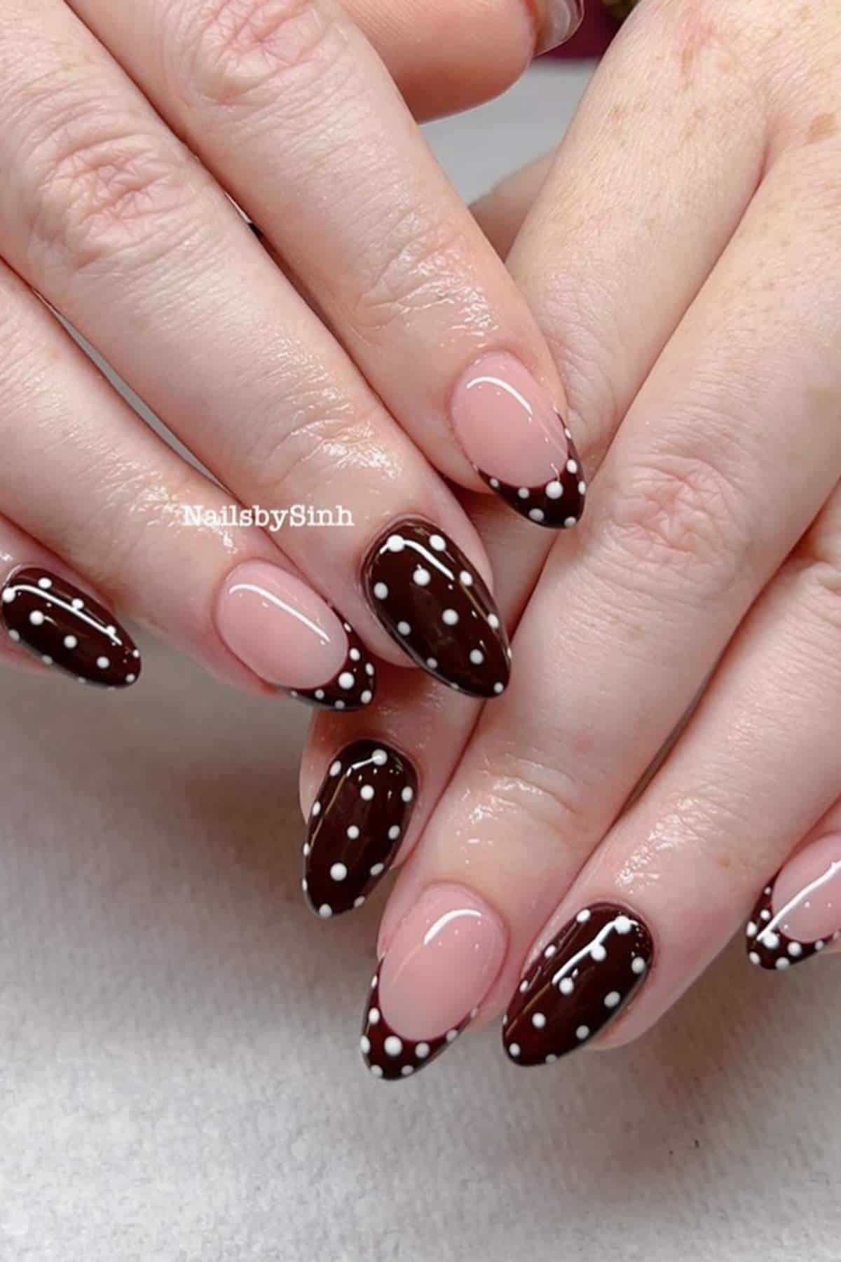 Hands with almond-shaped nails featuring a brown base and white polka dots on some nails, and French tips with brown and white polka dots on others.
