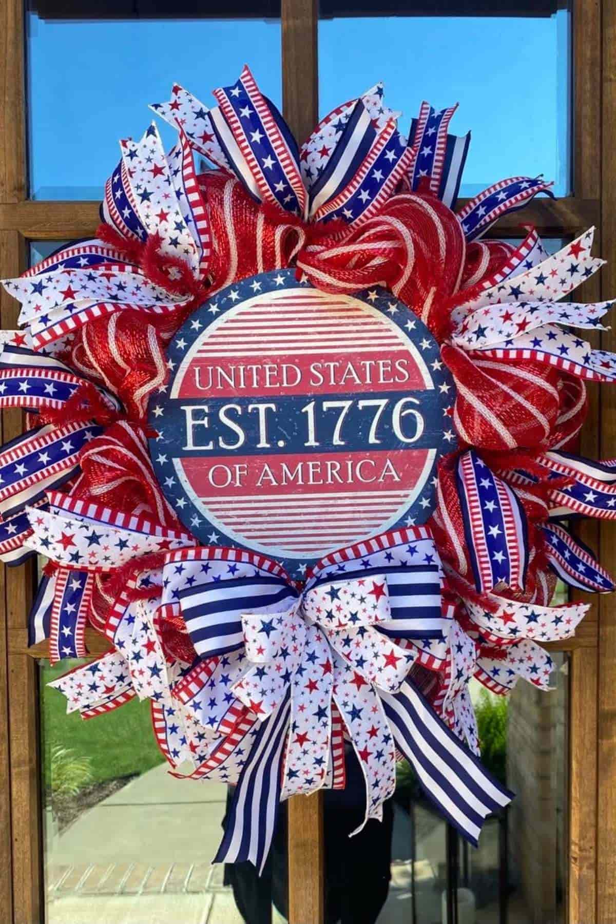 A wreath decorated with red, white, and blue ribbons and stars, featuring a sign that reads "United States of America EST. 1776.