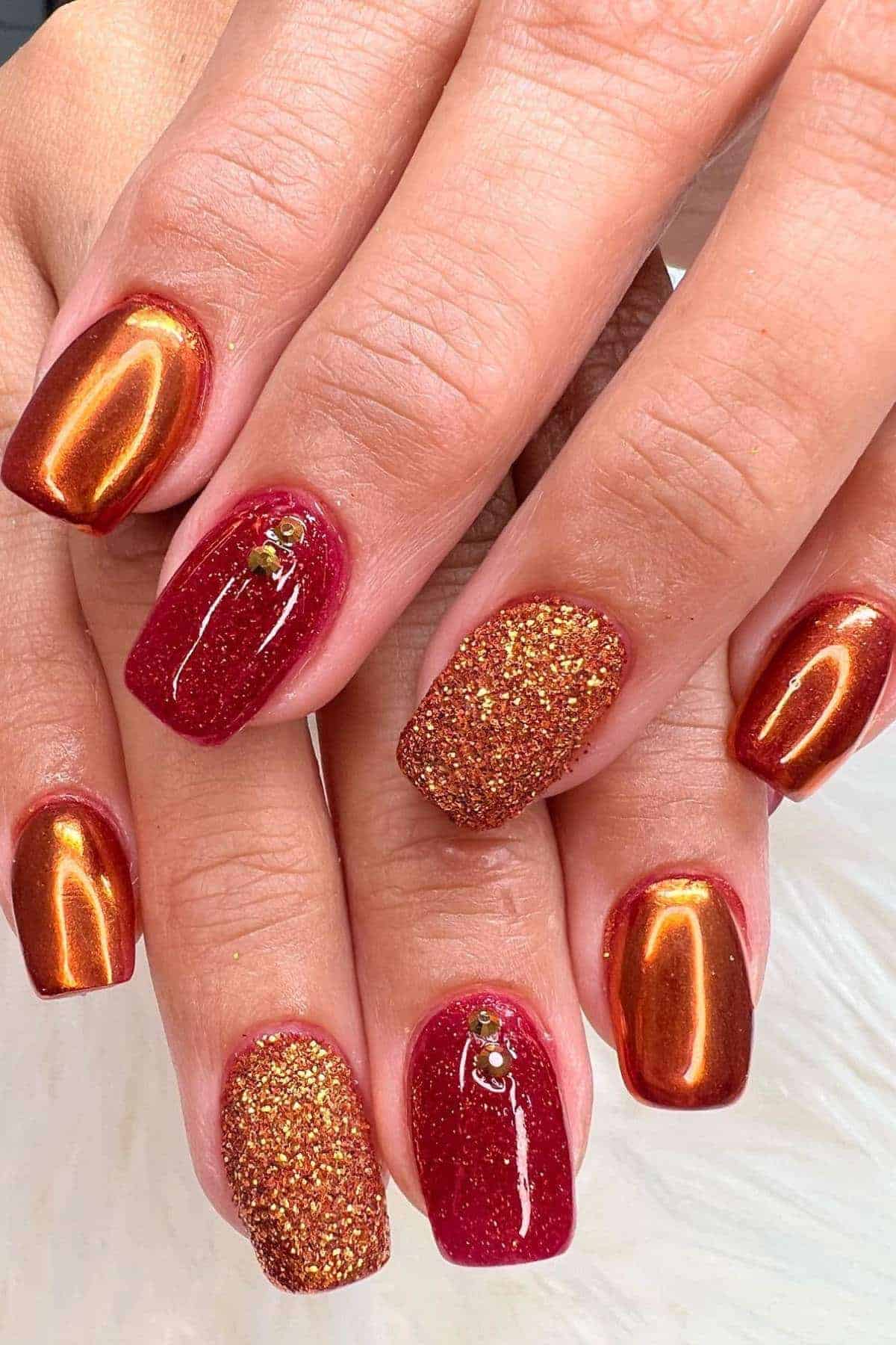 Close-up of hands with square-shaped nails featuring metallic copper, glittery gold, and deep red polish, some accented with small rhinestones.