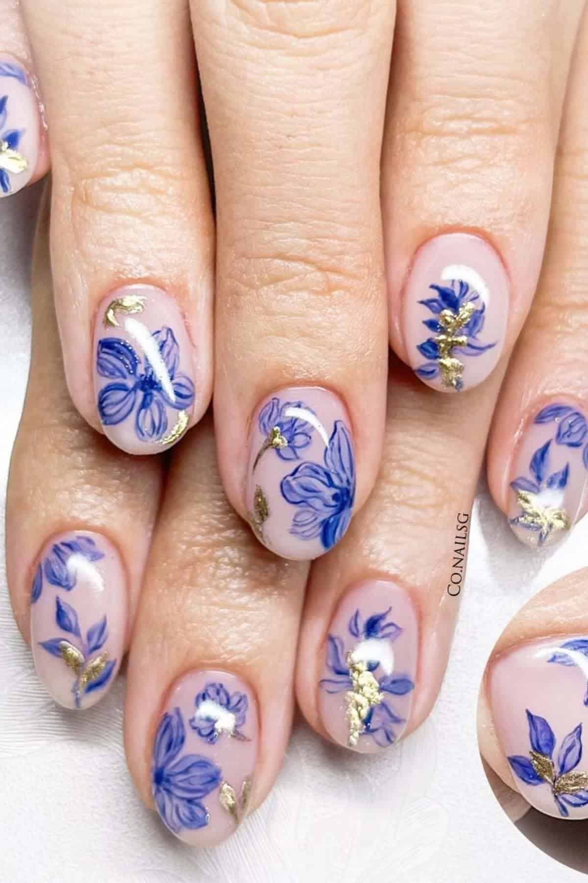 Close-up of fingernails with a light pink base, blue floral designs, and gold foil accents.