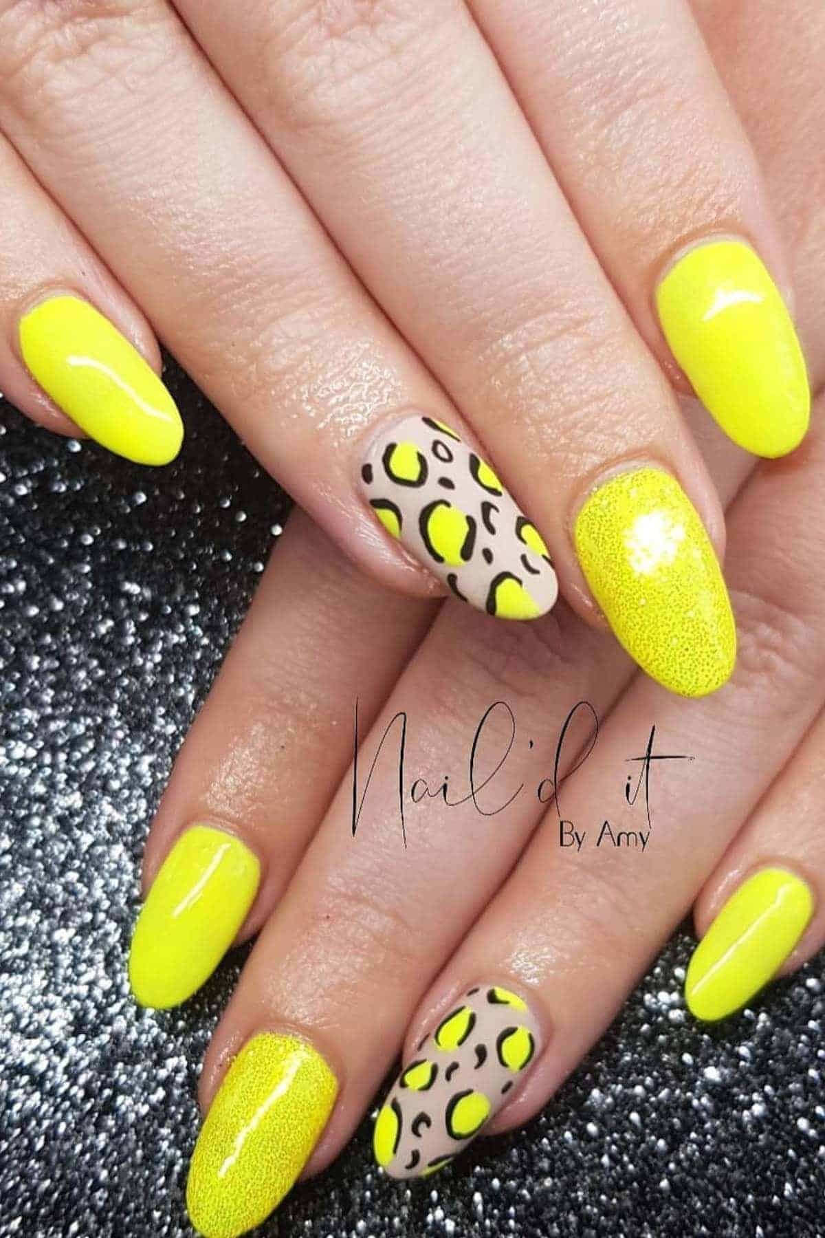 Hands with almond-shaped nails featuring bright yellow polish and two accent nails with a beige base and yellow and black abstract leopard print design.