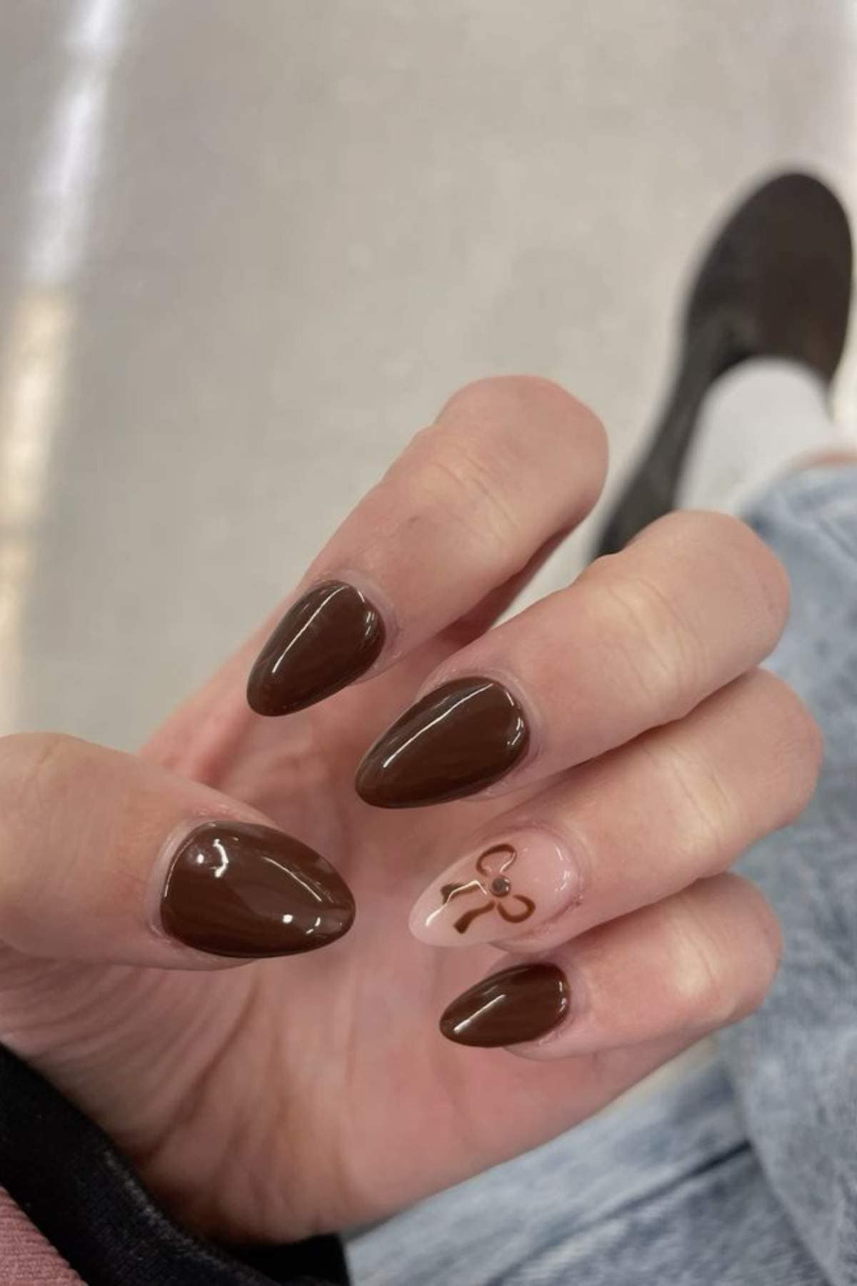 A hand with almond-shaped nails painted brown, except the ring finger which is nude with a small brown bow design.
