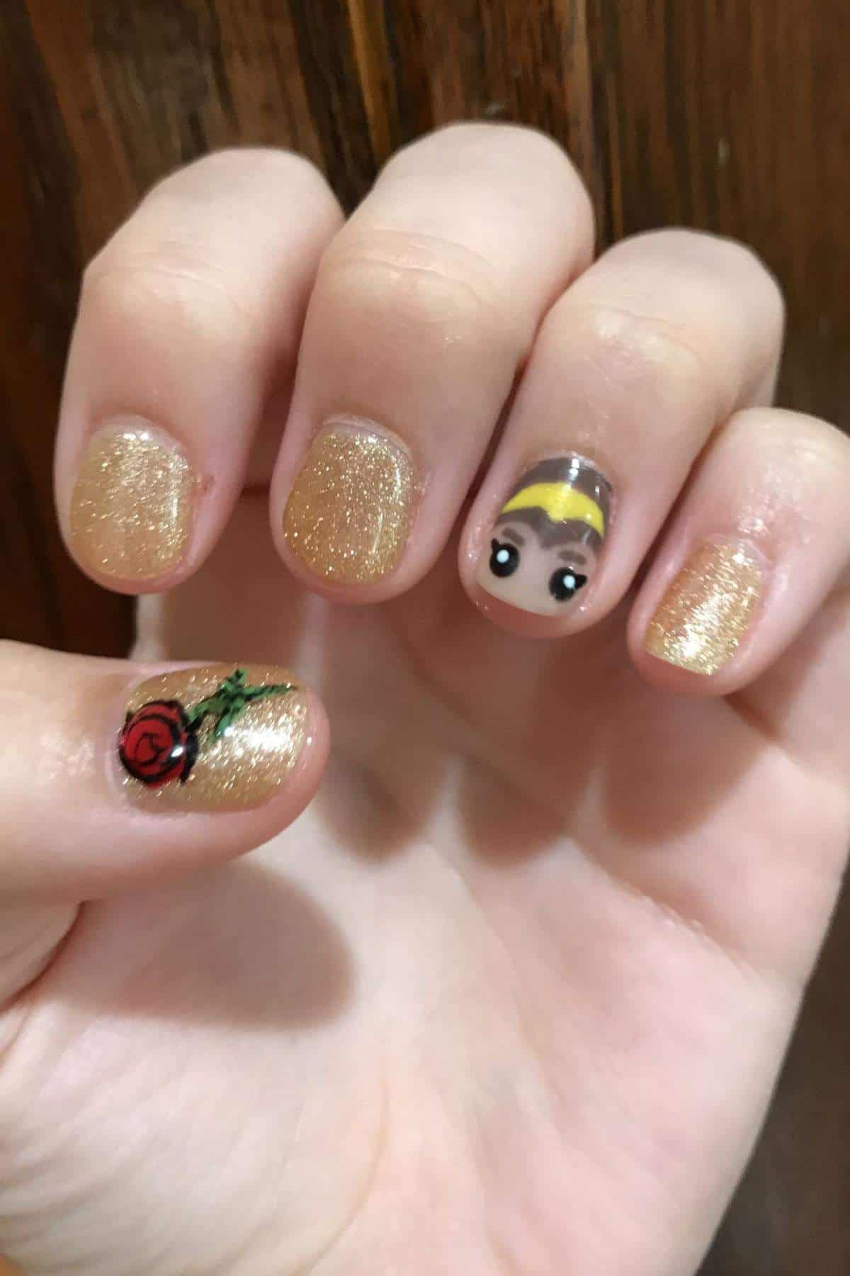 A hand with gold glitter nail polish; the thumb features a painted red rose, and the ring finger shows a cartoon face with brown hair and a yellow headband.