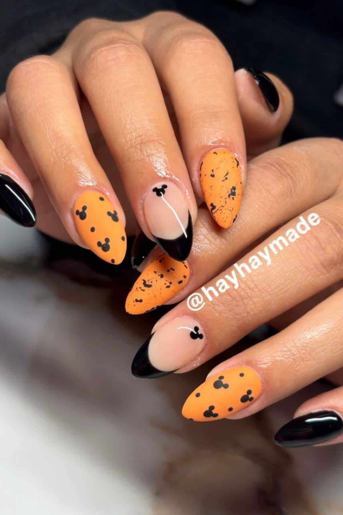 Hands with almond-shaped nails featuring a Halloween-themed manicure in orange, black, and nude, with Mickey Mouse silhouettes and black tips, tagged @hayhaymade.
