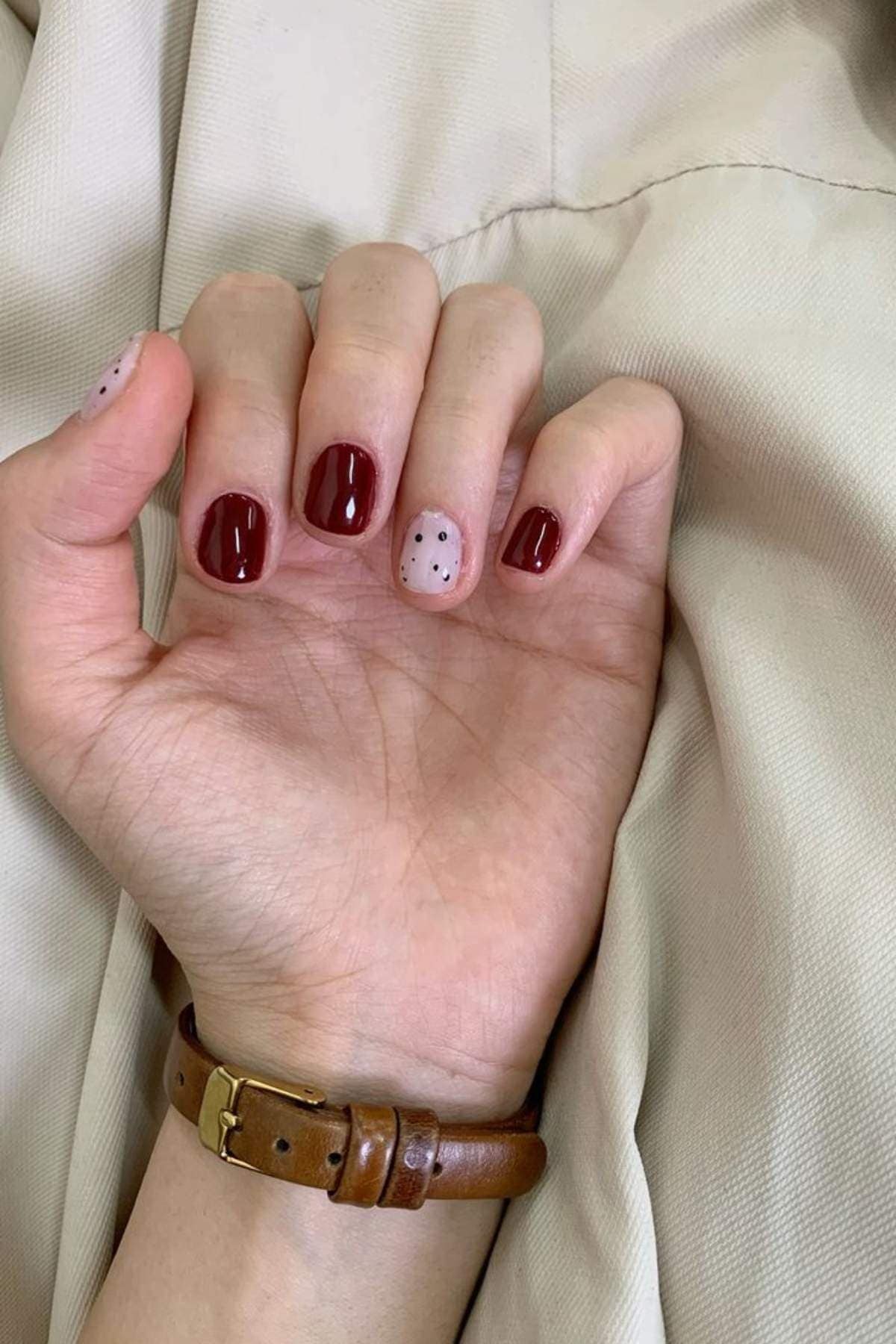 A hand with short nails, four painted dark red and one painted beige with black dots, resting on a cream-colored fabric. The wrist has a brown leather strap watch.