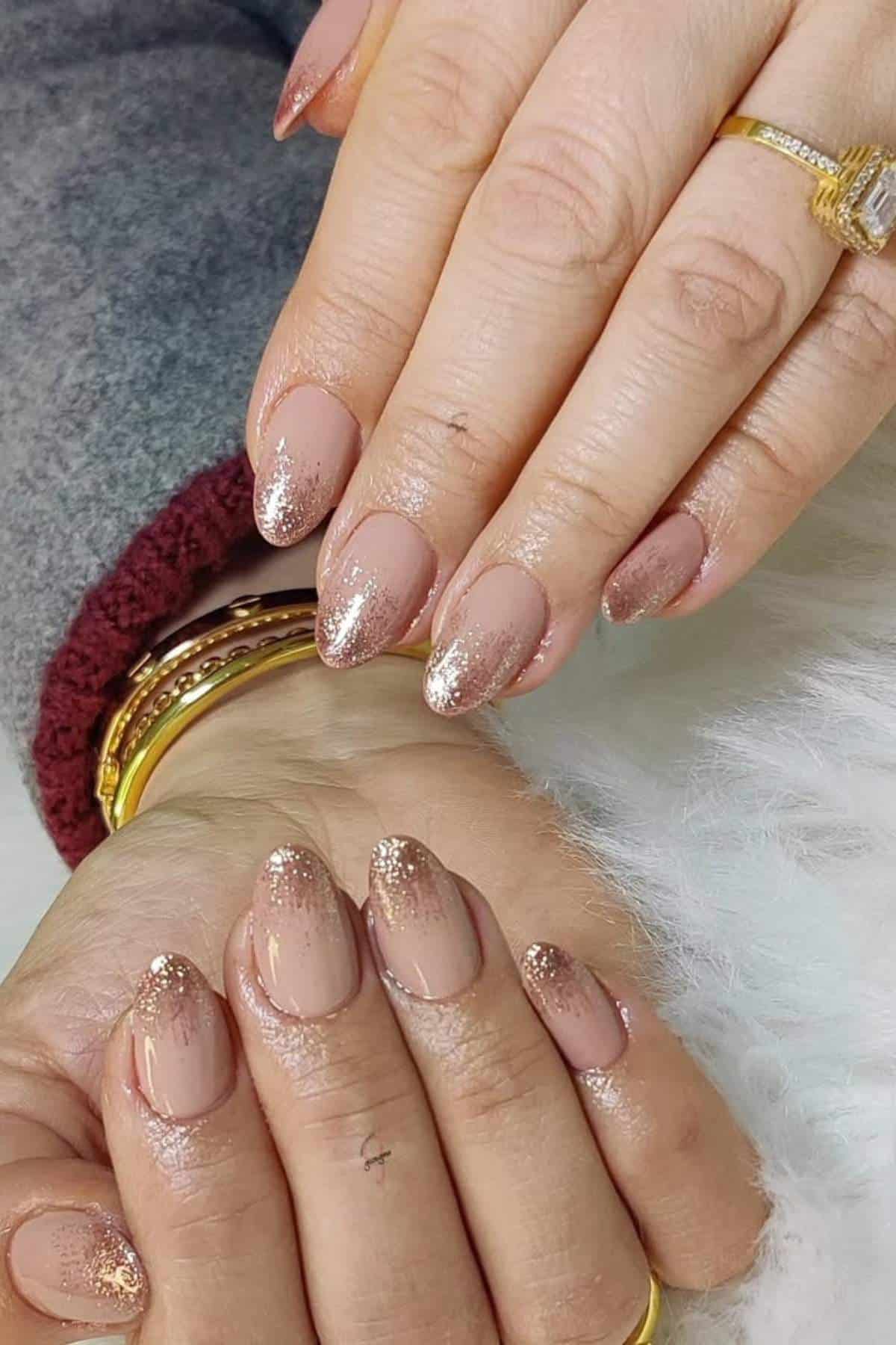 Hands with nude almond-shaped nails featuring gold glitter tips, wearing gold rings and bangles, resting on a white surface.