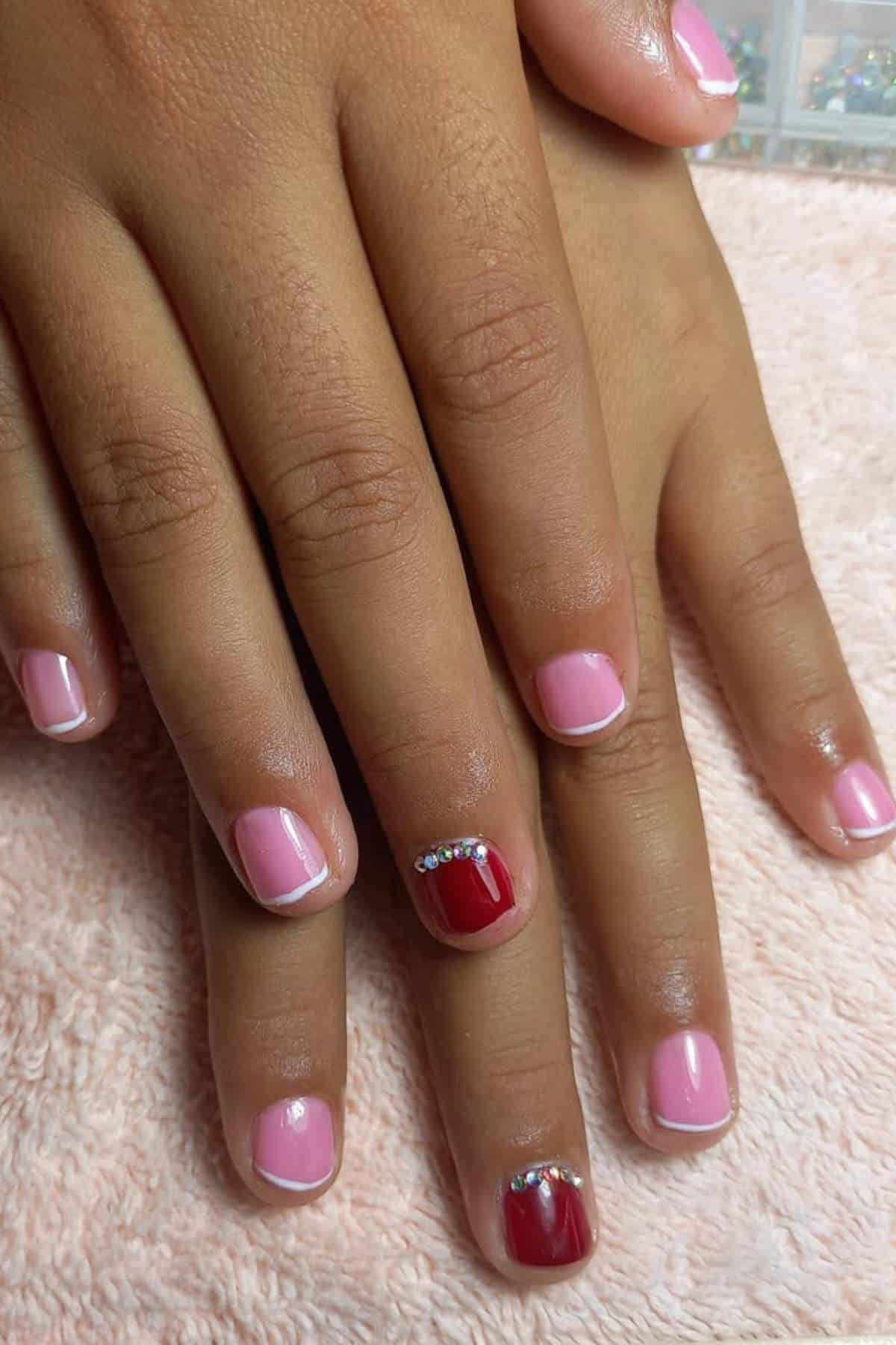 Close-up of hands with short, square-shaped nails painted pink; ring fingernails are painted red with small rhinestone decorations near the cuticles.