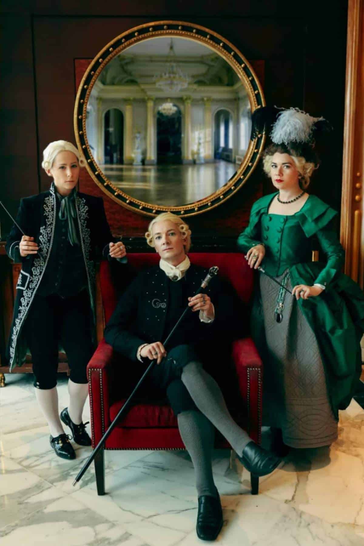 Three people in elaborate 18th-century costumes pose indoors; one sits on a red chair while the other two stand with canes, with a large round mirror behind them.