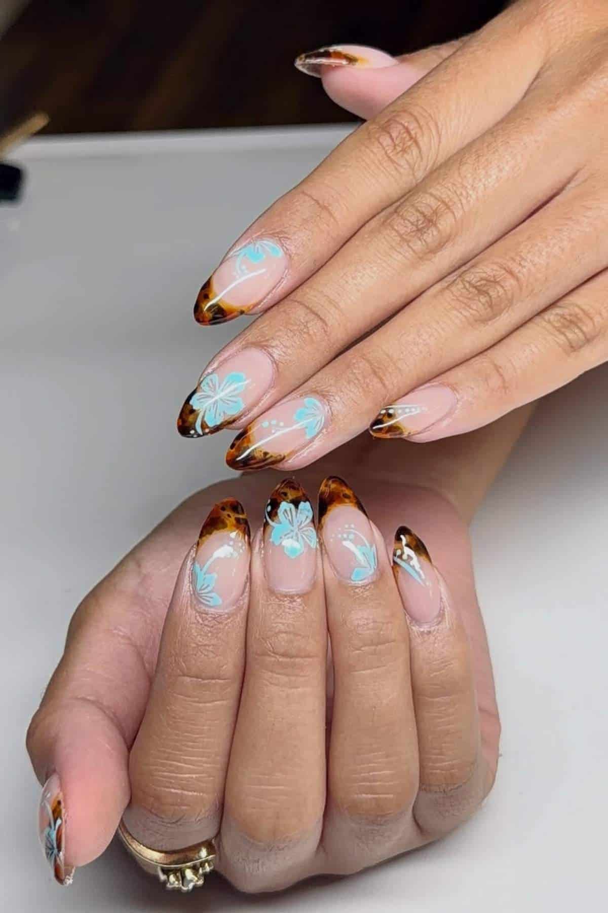 Hands with almond-shaped nails featuring tortoiseshell tips and light blue floral designs on a neutral base, resting on a white surface.
