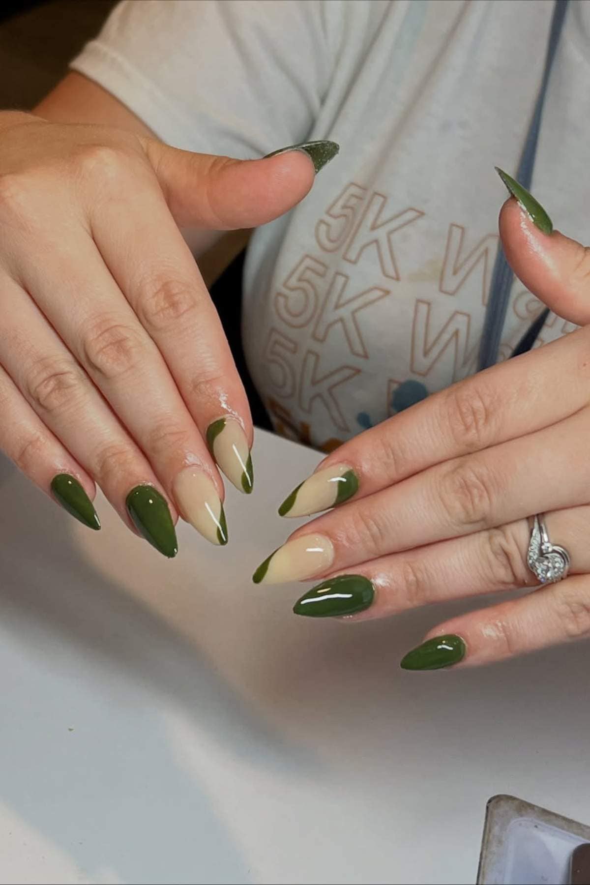 Two hands with almond-shaped nails painted in glossy olive green and beige, featuring alternating solid and diagonal split designs. A diamond ring is worn on the left hand.
