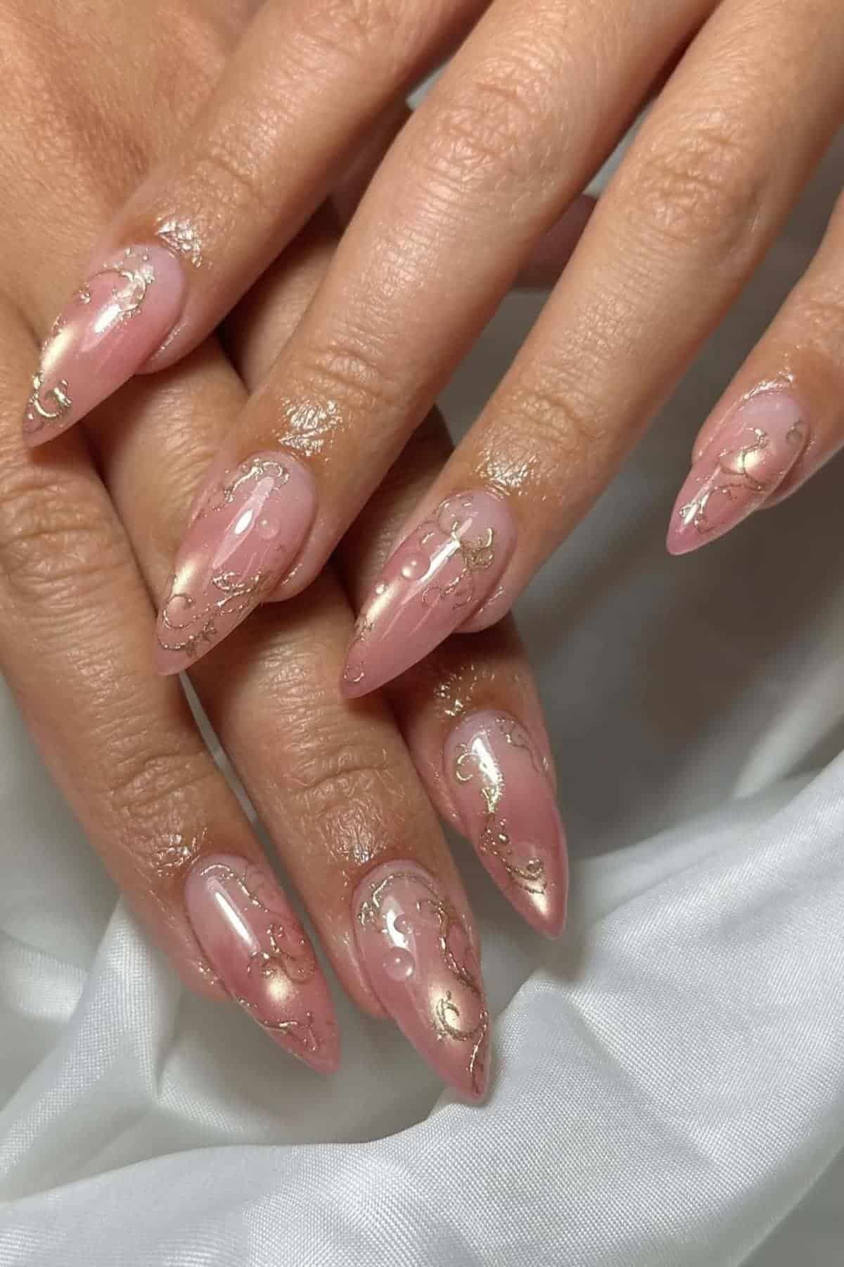 Hands with long, almond-shaped nails painted in a pink nude shade with gold foil accents and delicate swirl designs, resting on a white fabric background.