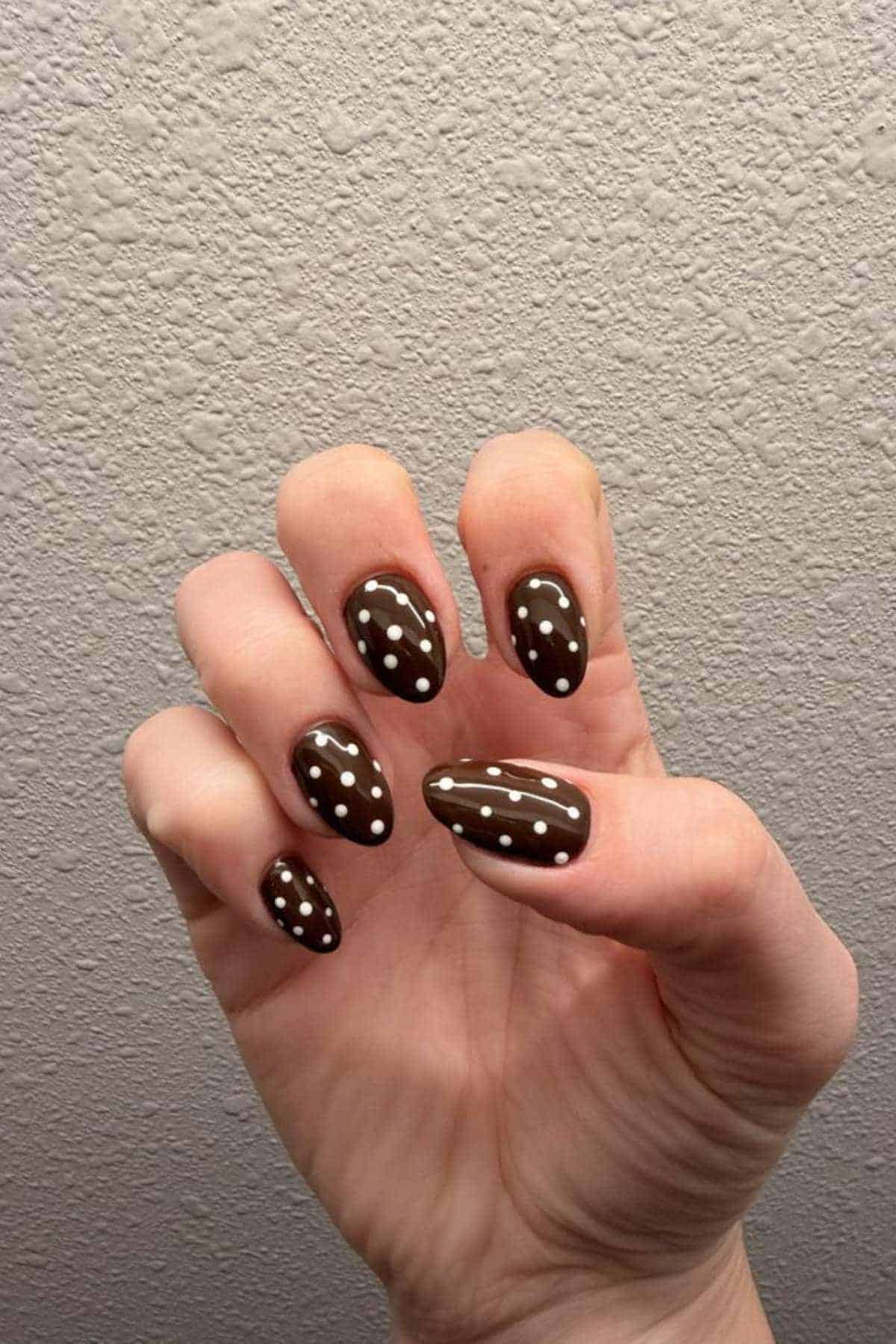 A hand with brown almond-shaped nails, each featuring white polka dot and line designs, held against a textured light-colored wall.