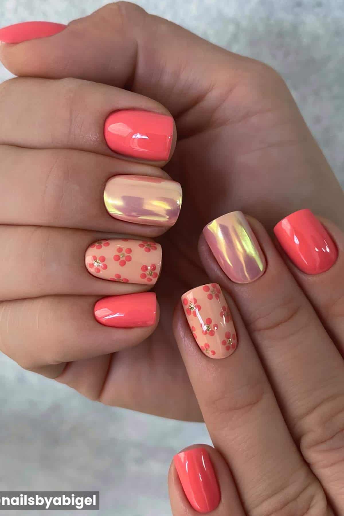 Hands with short, square-shaped nails painted in coral, nude, and iridescent shades, featuring small floral designs and a glossy finish.