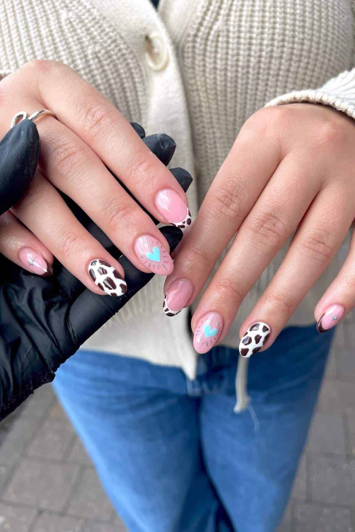 Hands with manicured nails featuring pink polish, blue leaf designs, and black-and-white animal print accents. Person is wearing a cream sweater and blue jeans.
