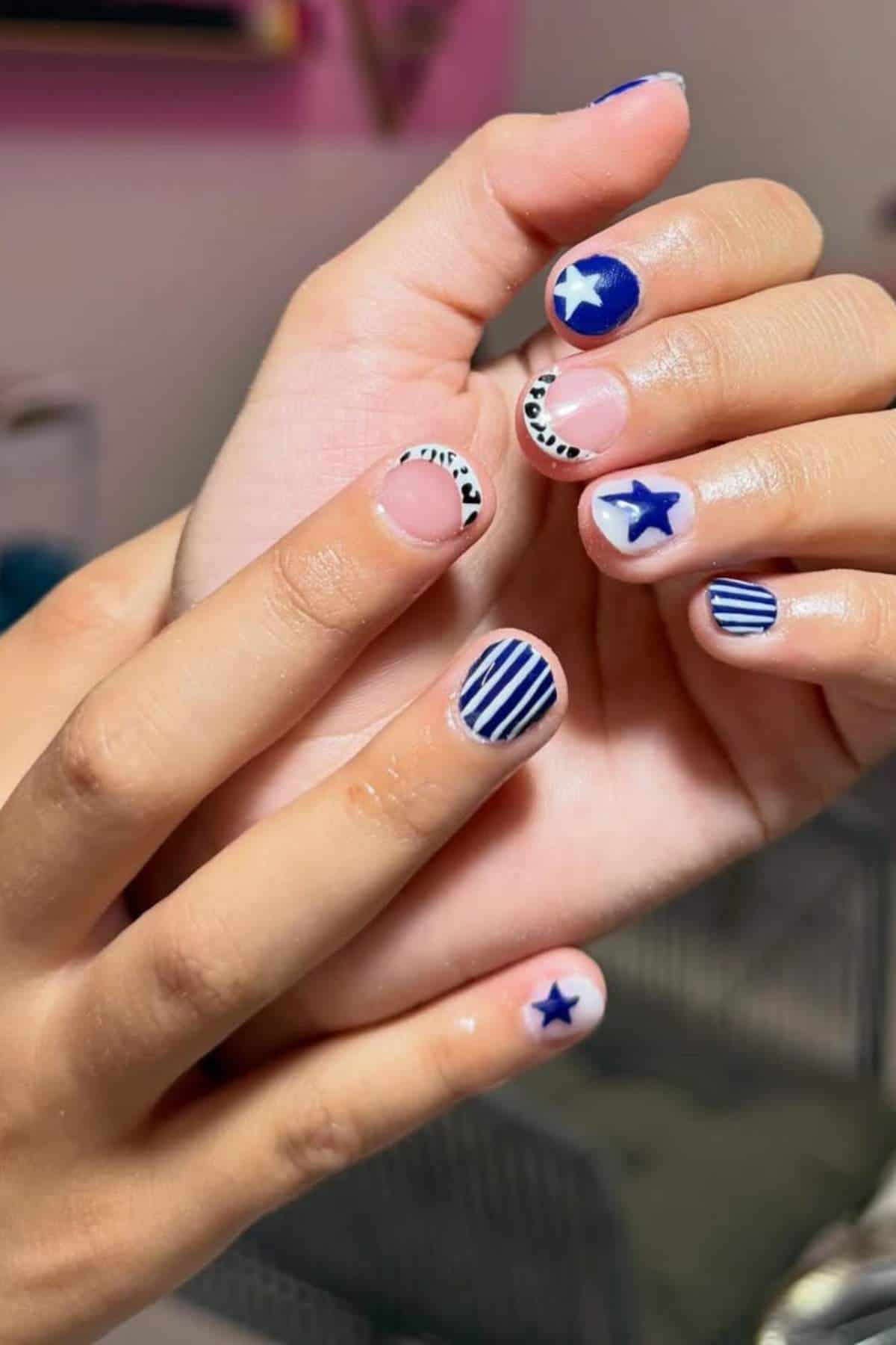 Close-up of hands with short nails painted in blue and white designs, including stripes, stars, and abstract patterns.