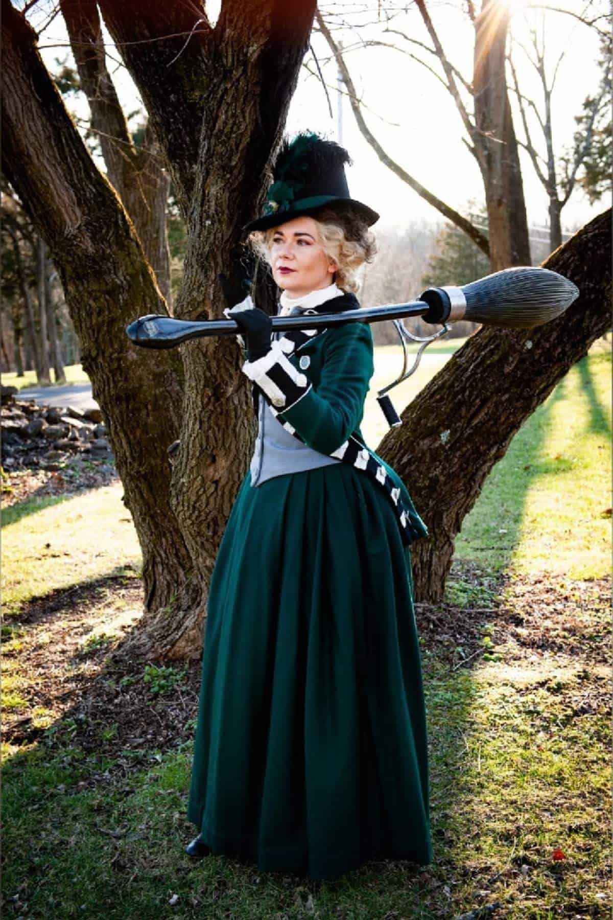 A person in a green Victorian-style dress and hat stands outdoors holding a broomstick over one shoulder, with trees and sunlight in the background.
