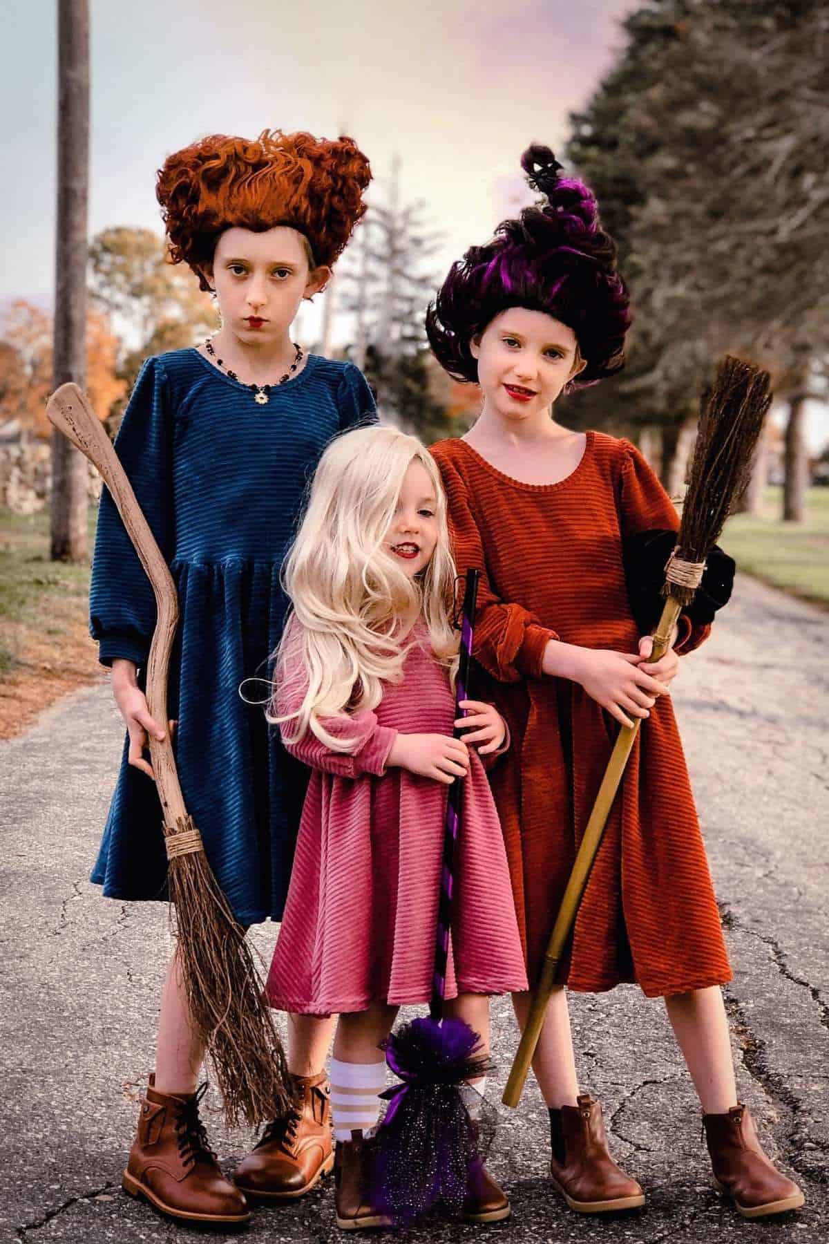 Three children in colorful dresses and wigs stand outdoors on a road, each holding a broom, dressed as witches.