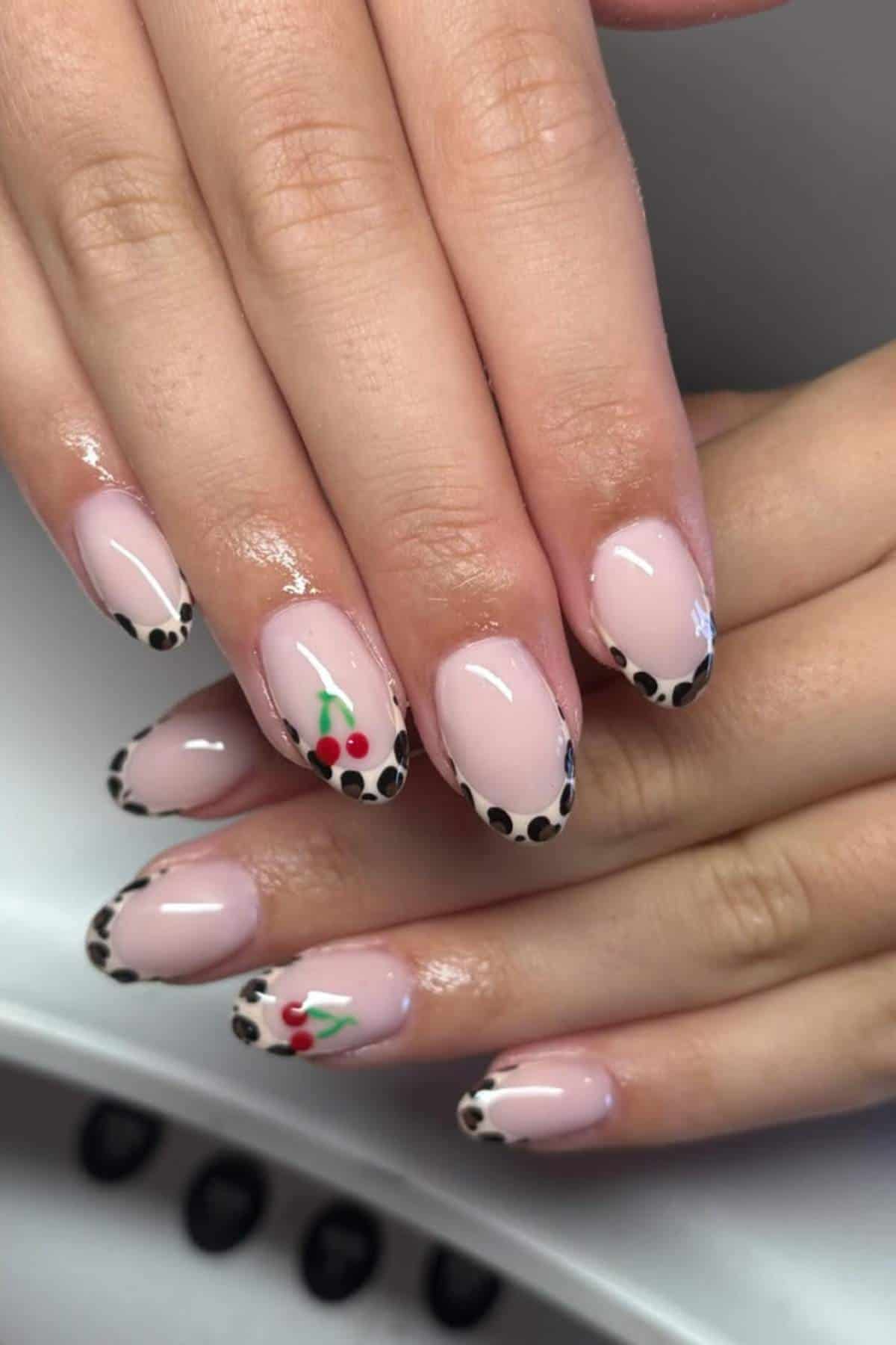 Hands with almond-shaped nails featuring pale pink polish, black and white polka dot French tips, and small cherry designs on two accent nails.