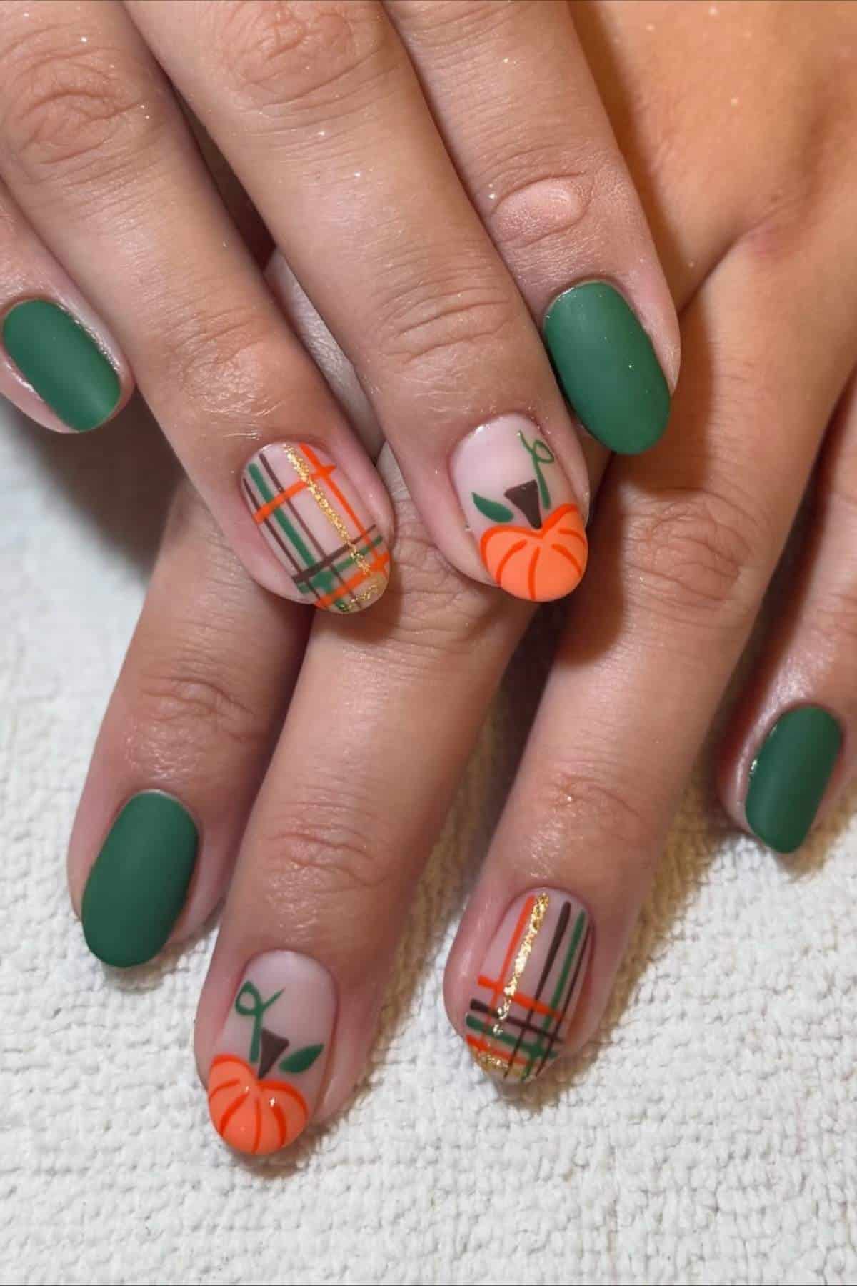 Hands with short nails painted matte green and accent nails featuring orange pumpkins and plaid designs in green, orange, and gold, resting on a white surface.