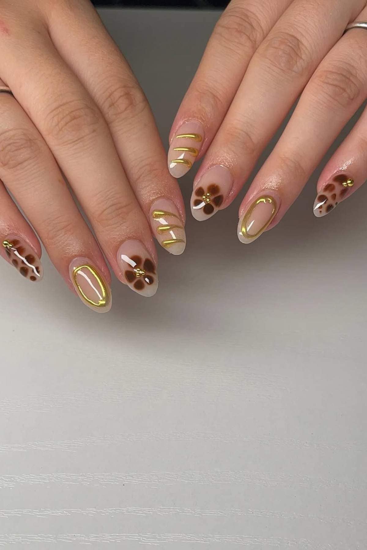 A set of manicured hands with almond-shaped nails featuring gold outlines, gold stripes, and brown flower designs on a light base.