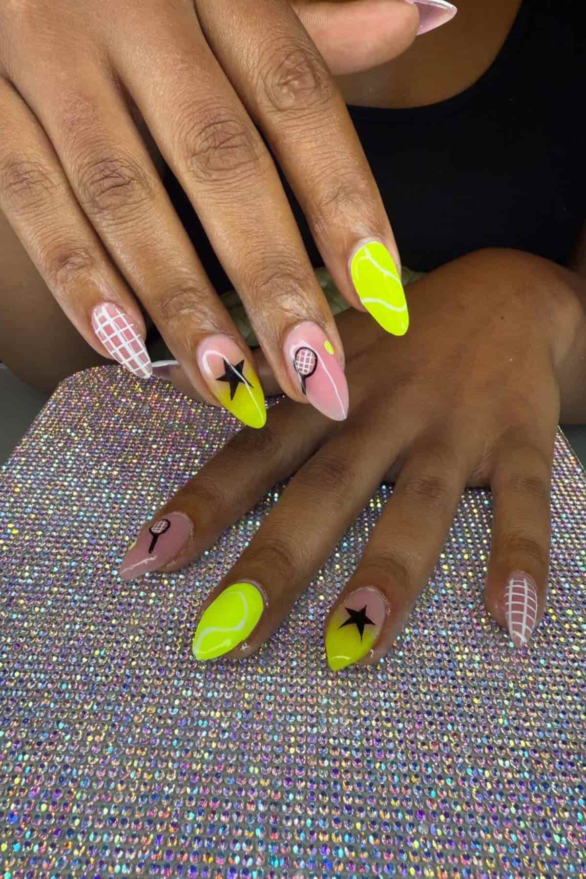 Hands with long, almond-shaped nails featuring tennis-themed nail art, including tennis balls, rackets, nets, and stars in yellow, pink, black, and white on a sparkly surface.