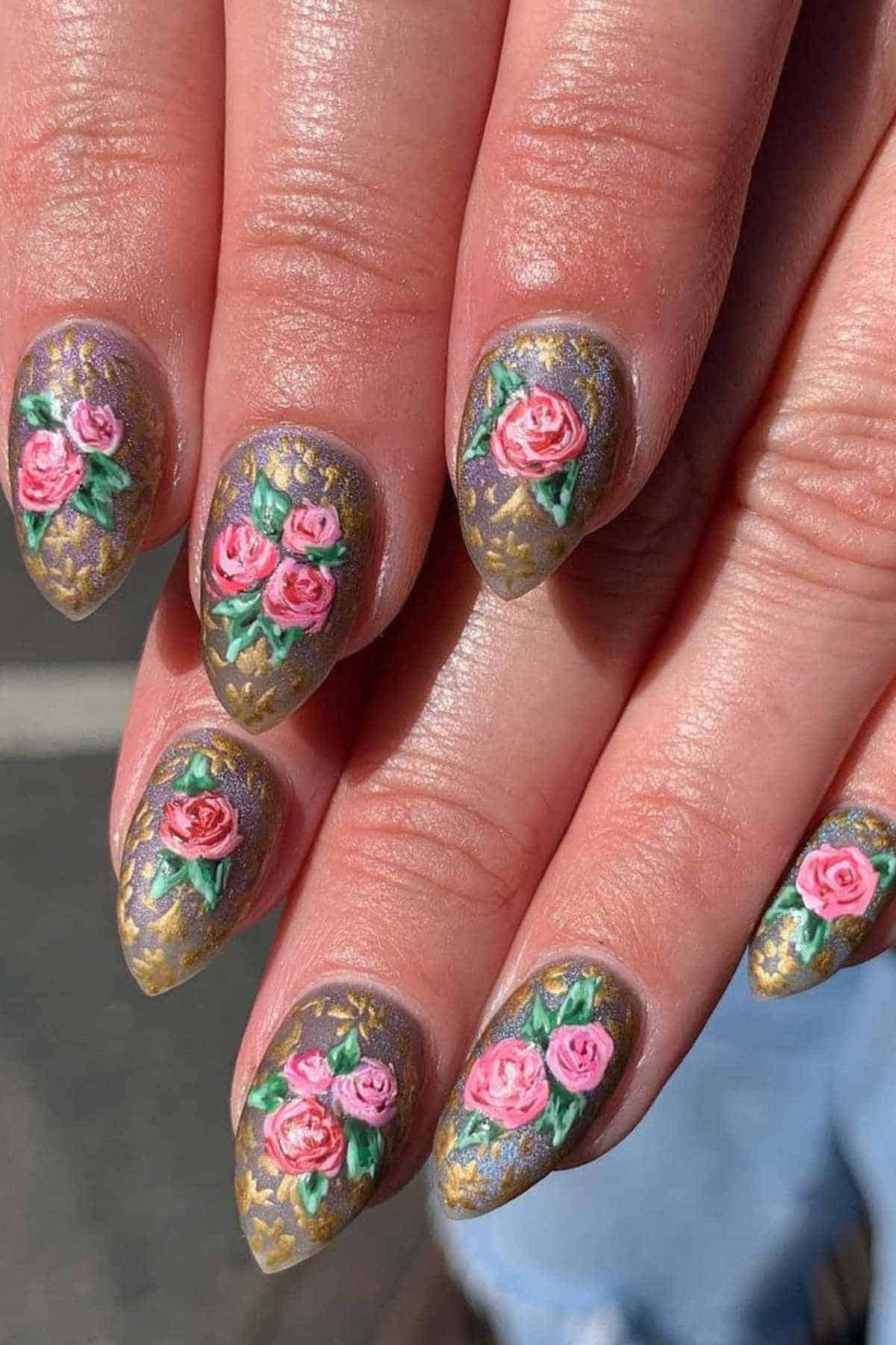 Close-up of hands with almond-shaped nails featuring 3D pink rose nail art, gold detailing, and a shimmery purple base.
