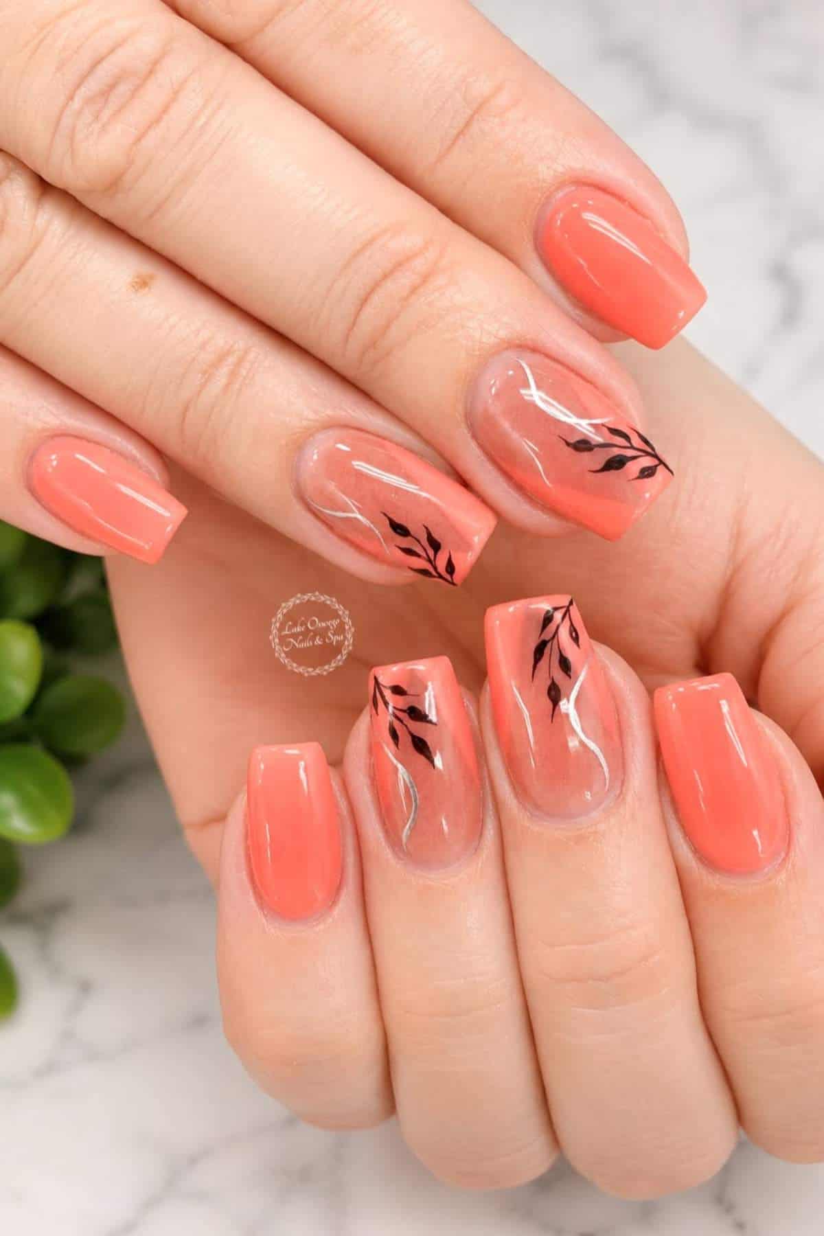 Hands with short, square-shaped coral nails; some nails have black leaf and white accent designs on a clear base. Marble surface and green plant in the background.