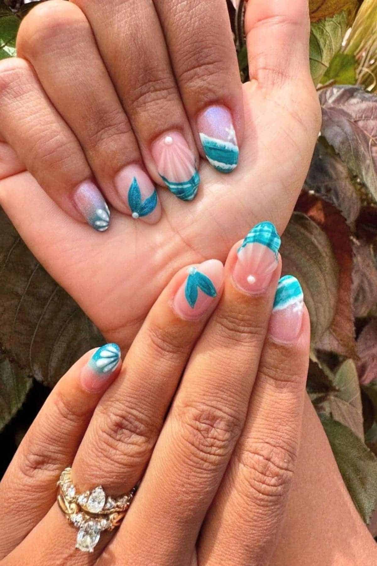 Hands with blue, turquoise, and white nail art featuring leaf, wave, and shell designs. One hand has three gold rings with clear stones. Background includes green and brown leaves.