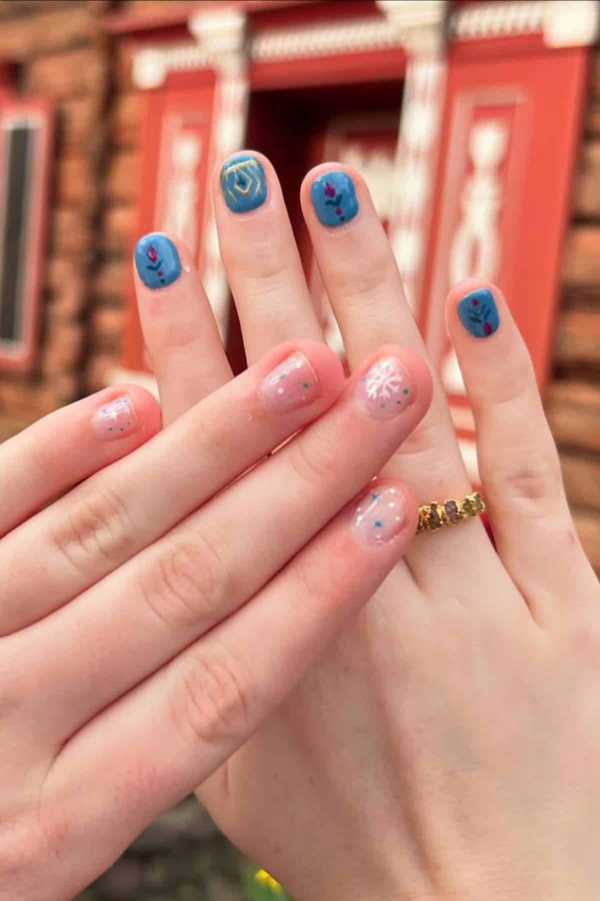 Two hands with manicured nails, one with blue polish and flower designs, the other with light pink polish and snowflake designs, held in front of a wooden building with red trim.