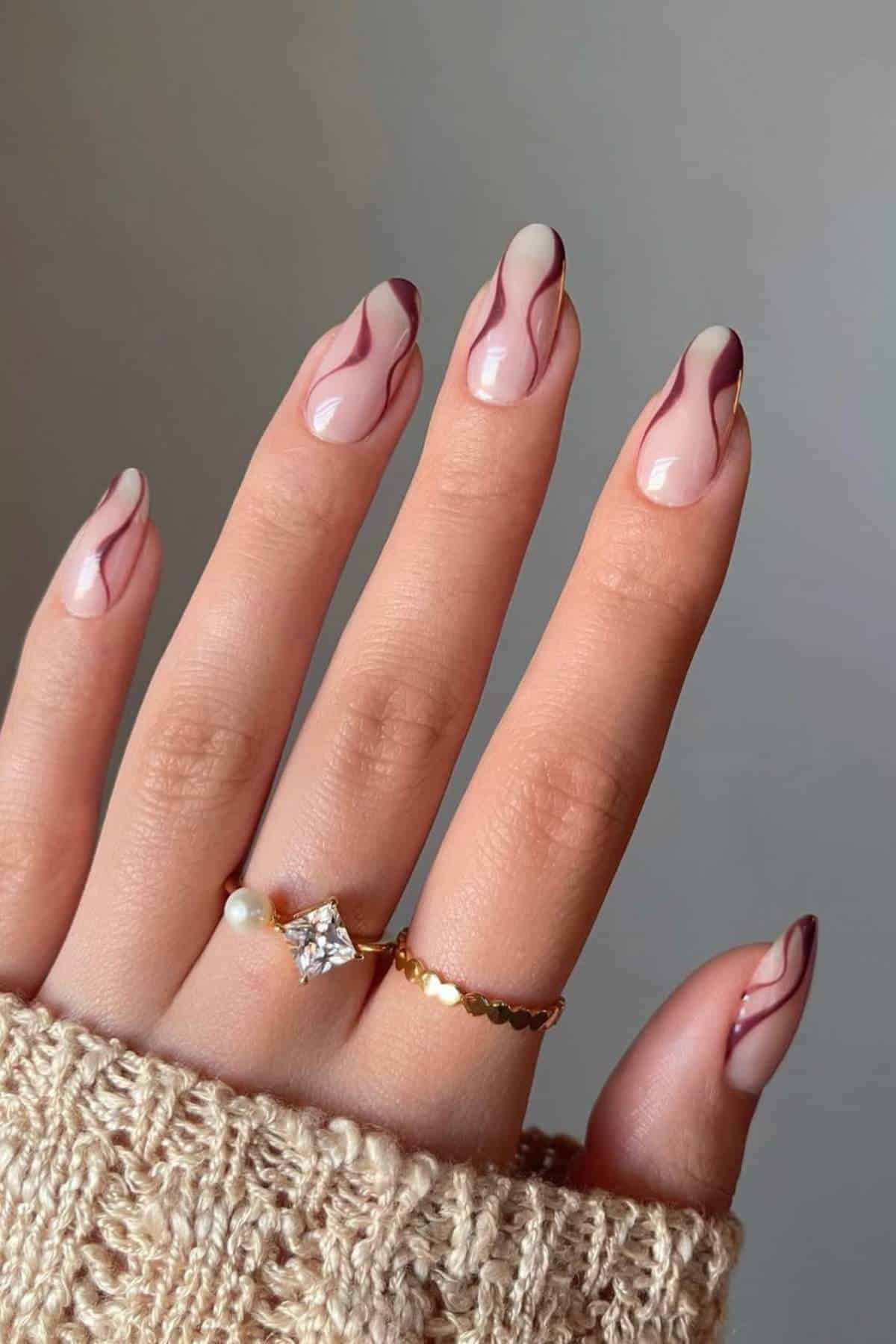 A hand with almond-shaped nails features a nude base and abstract dark brown wave designs; the person wears two rings and a beige knit sleeve.