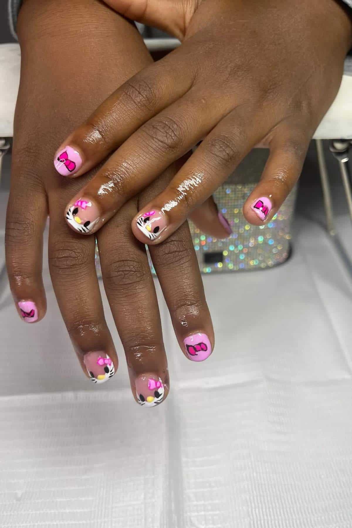 Close-up of hands with pink nail polish and cartoon cat designs, featuring black and white accents and some glitter on a few nails.