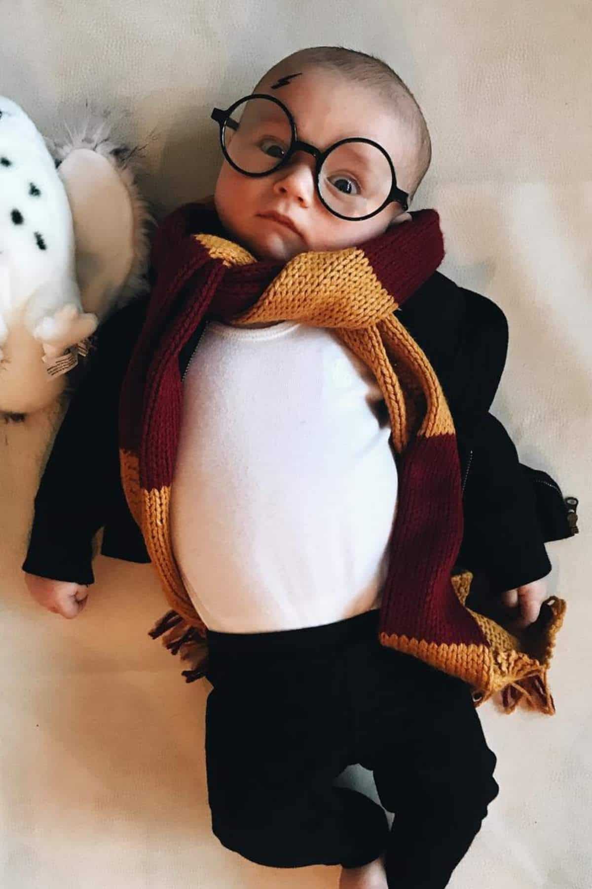 Baby dressed as Harry Potter with round glasses, a lightning bolt scar on the forehead, a maroon and yellow scarf, black jacket, and white shirt, lying on a light surface.