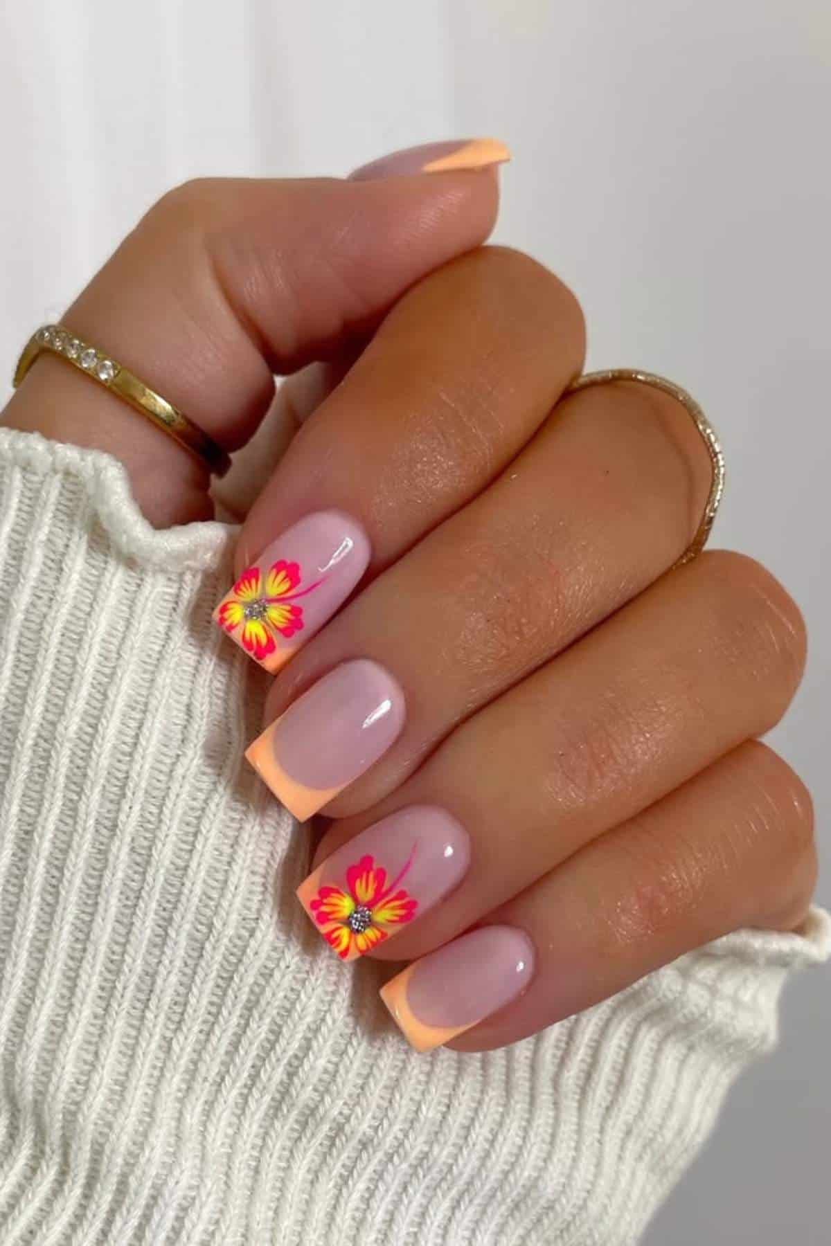 A hand with peach-colored French tip nails, featuring yellow and red flower designs on the ring and middle fingernails. The person wears two gold rings and a white ribbed sweater.