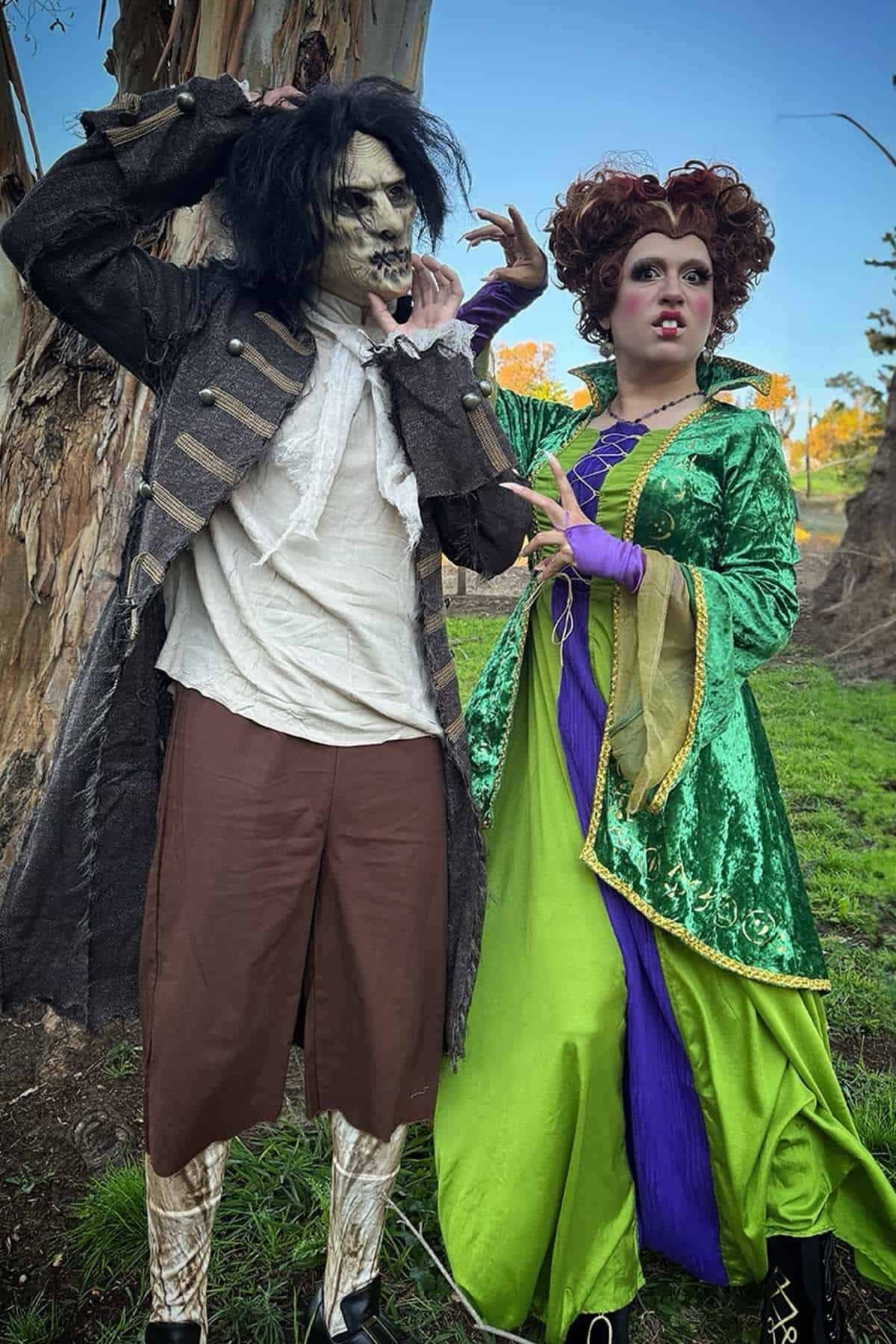 Two people in elaborate costumes stand outdoors; one wears tattered clothing and a monster mask, the other wears a green and purple dress with dramatic makeup and a curly wig.