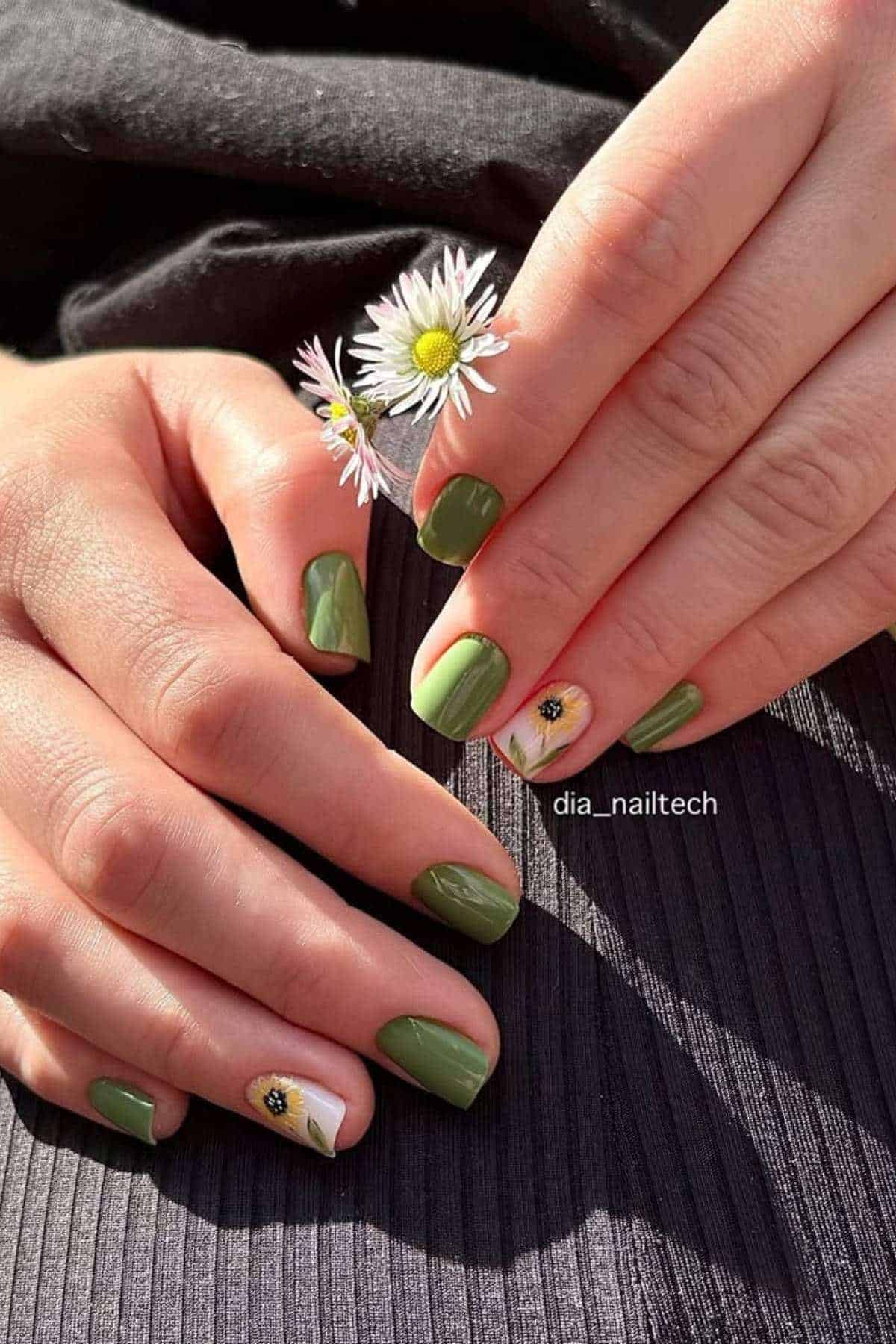 Hands with short green and olive nails, one accent nail featuring a daisy flower design, holding a small daisy flower.