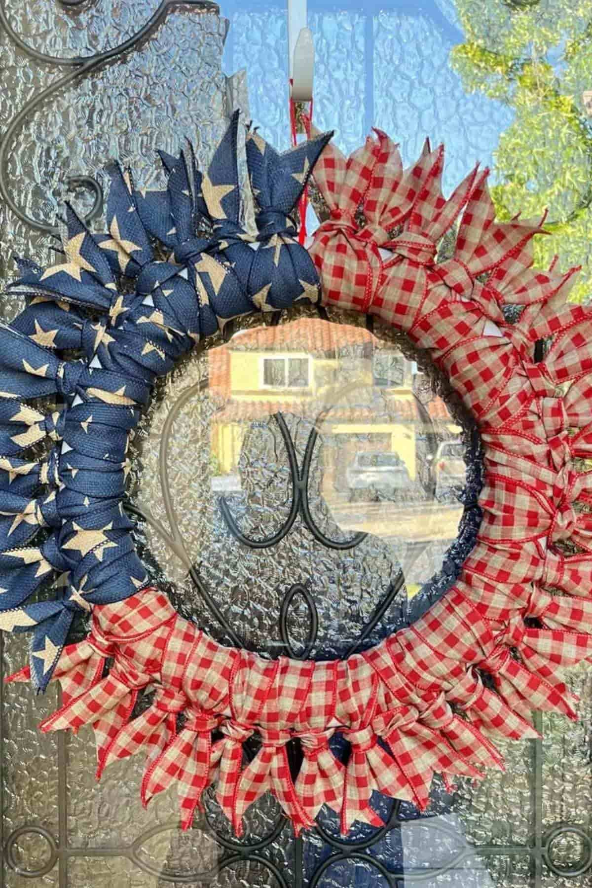 A wreath decorated with blue fabric featuring white stars on one side and red-and-white checkered fabric on the other, hanging on a glass door.