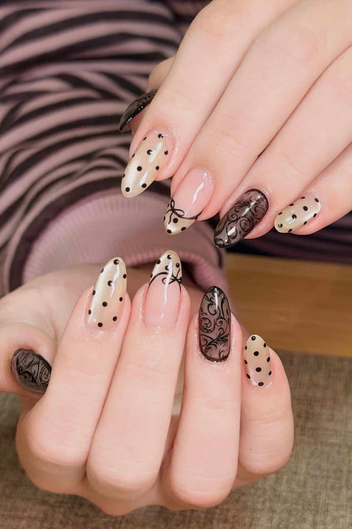 Hands with almond-shaped nails featuring a mix of beige and black polish, polka dots, and intricate black floral designs on a striped shirt background.