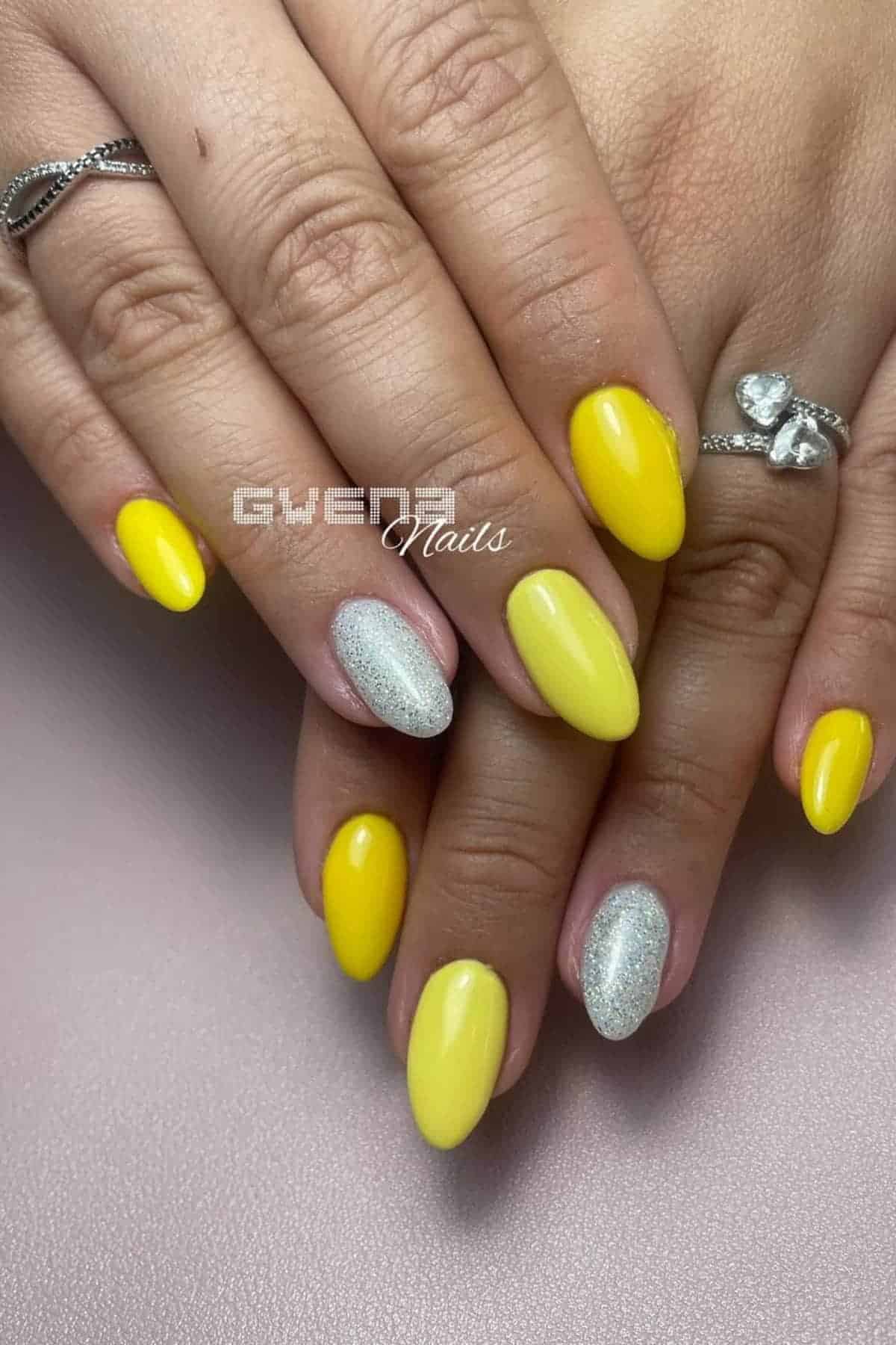Hands with almond-shaped nails featuring bright yellow polish on most nails and silver glitter polish on the ring fingers. The person is wearing silver rings.