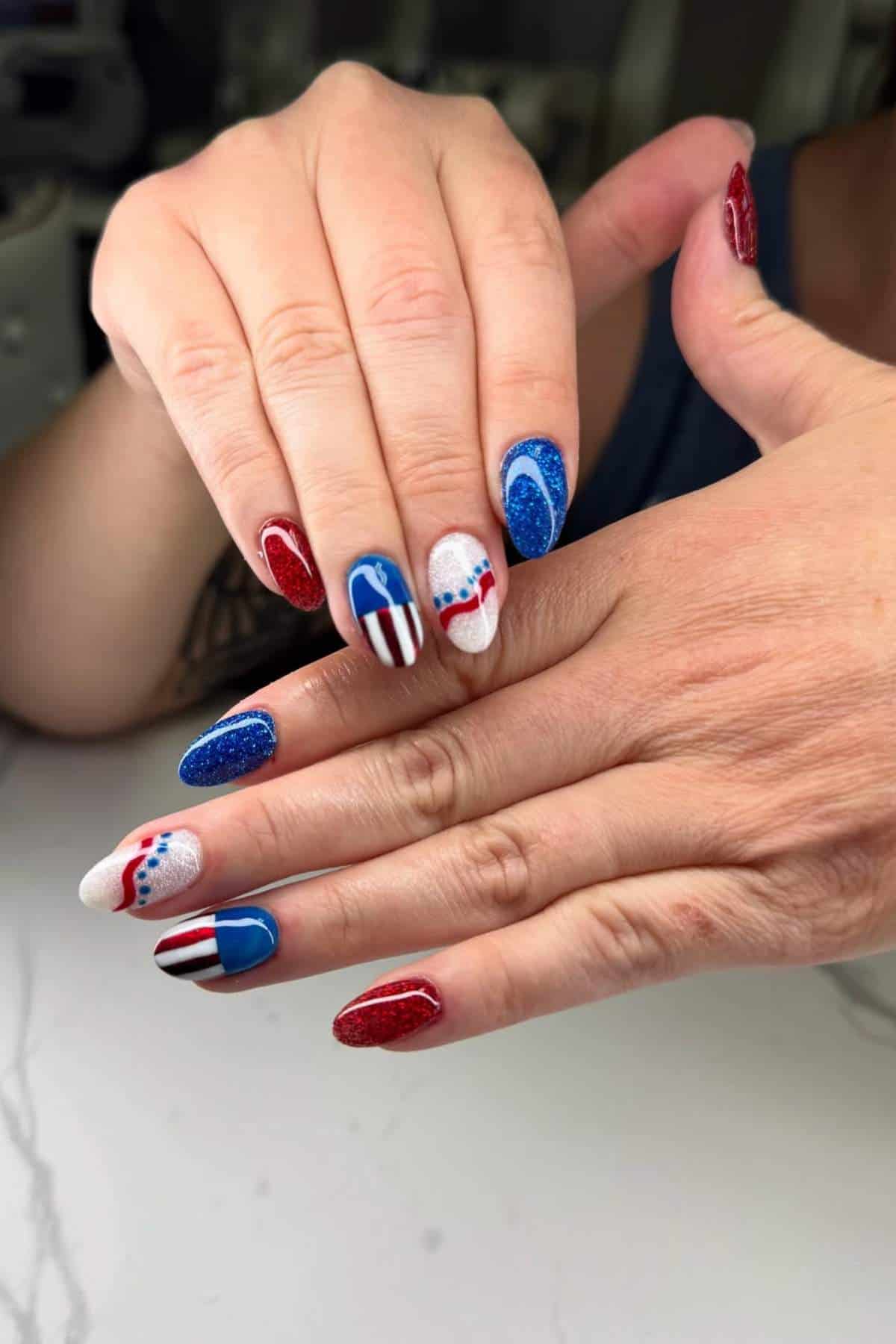 Hands with manicured nails painted in red, white, and blue with various patriotic designs, including stripes, stars, and dots.
